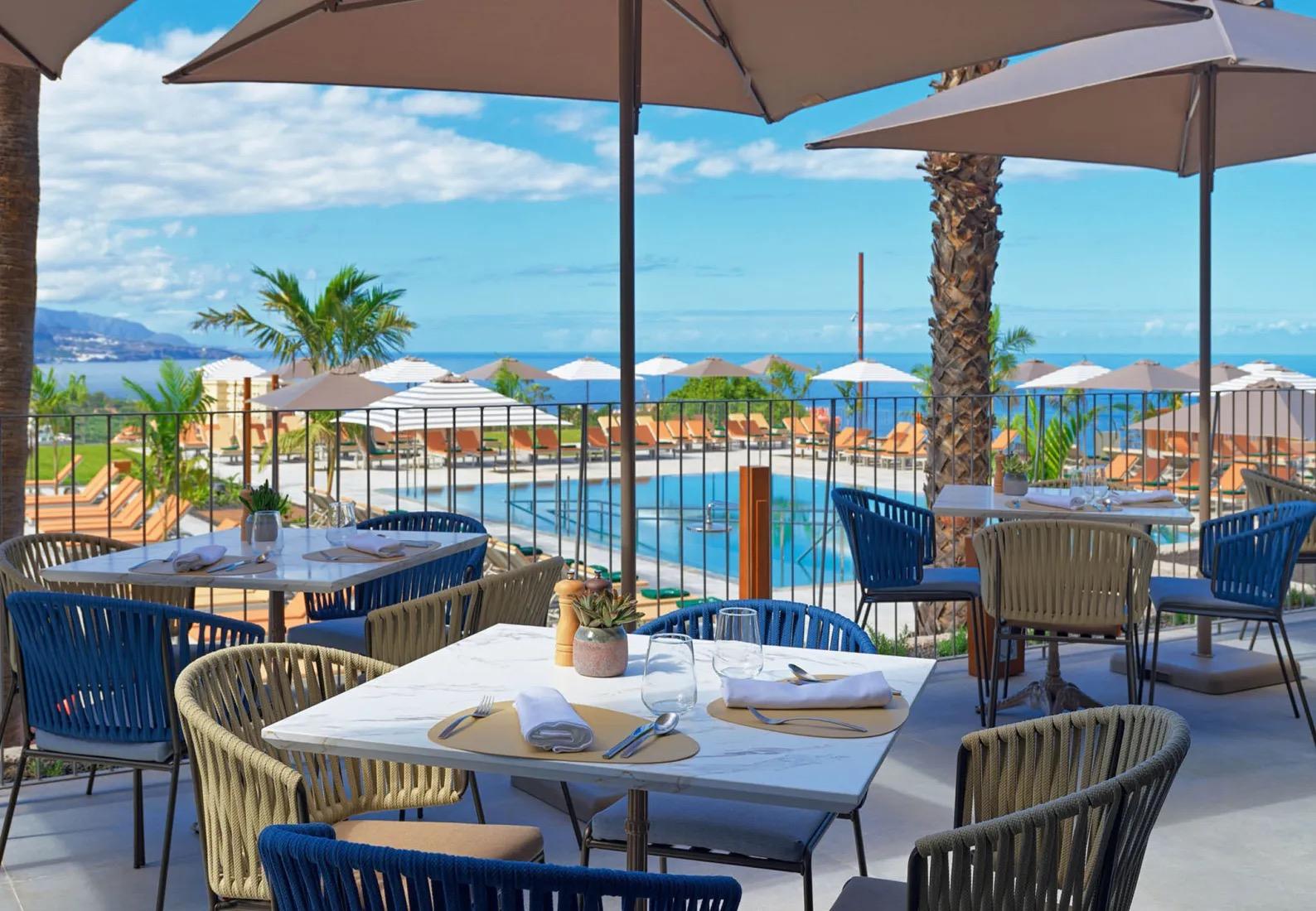 A restaurant terrace with marble tables set for lunch under umbrellas, looking over a swimming pool with loungers to the sea beyond.