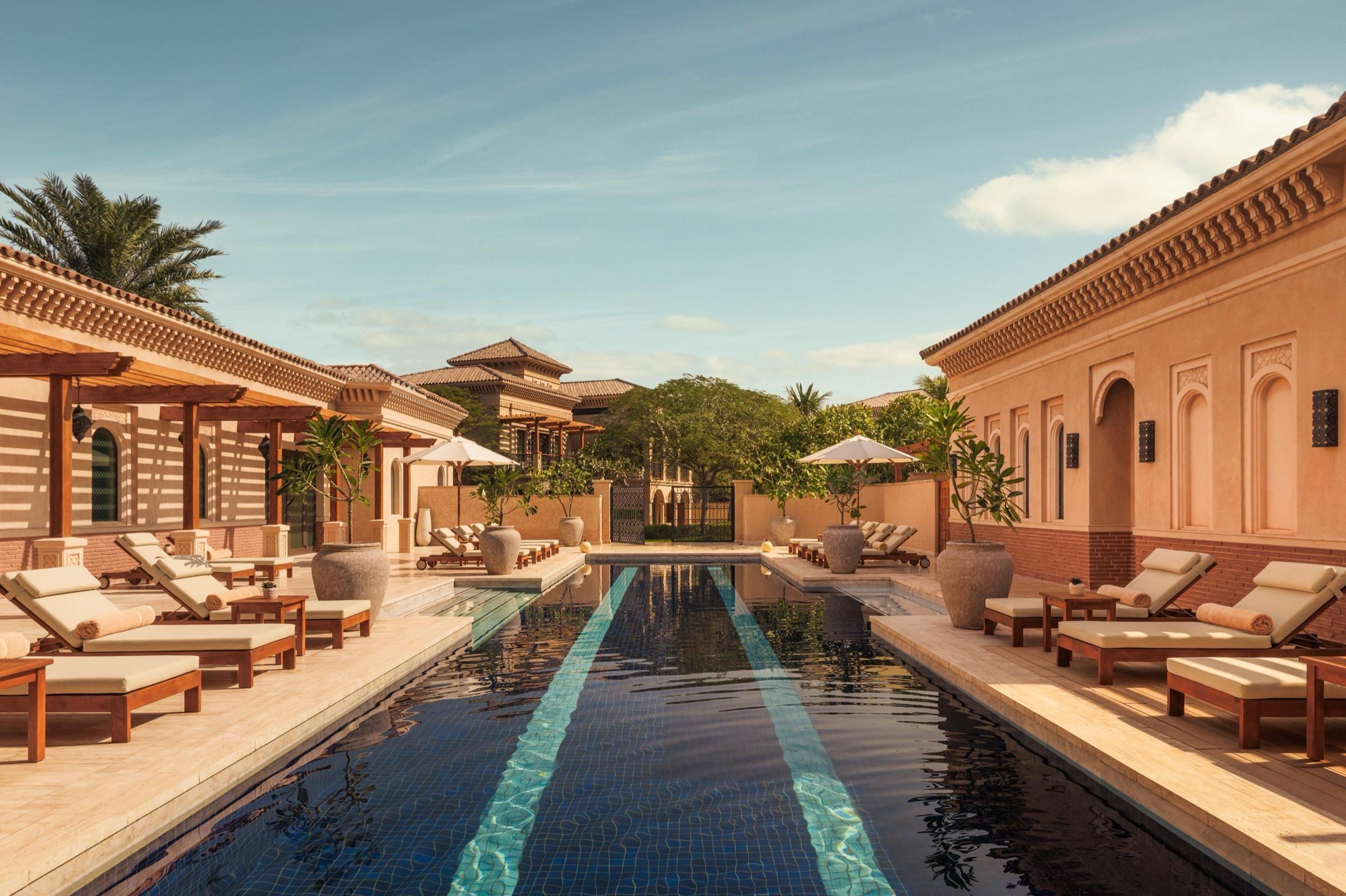 A narrow pool with a blue mosaic stripe runs between two rows of loungers and potted plants, flanked by sand-colored buildings.