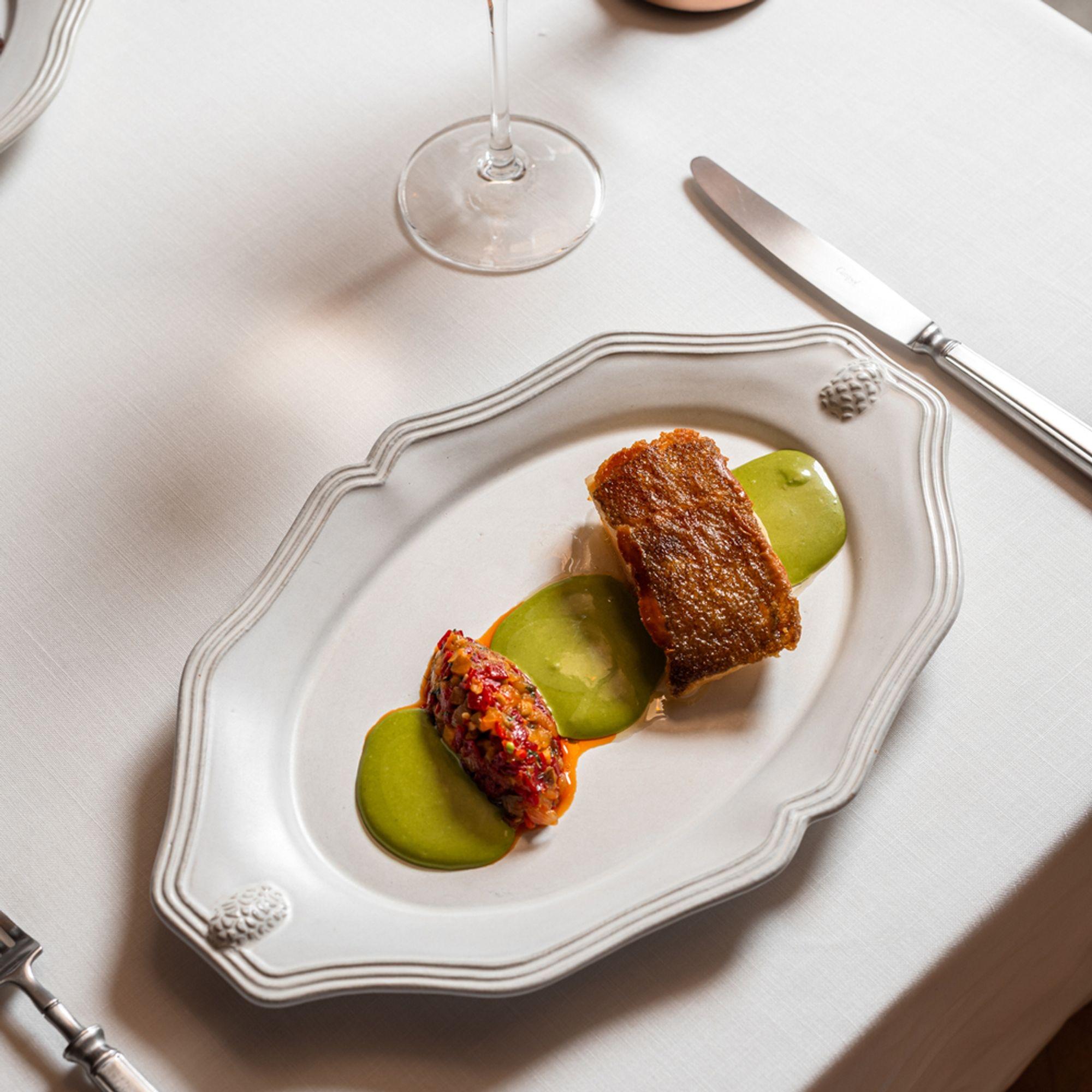 A plated dish of glazed meat on green purée on a scalloped white dish, a knife and wine glass beside it