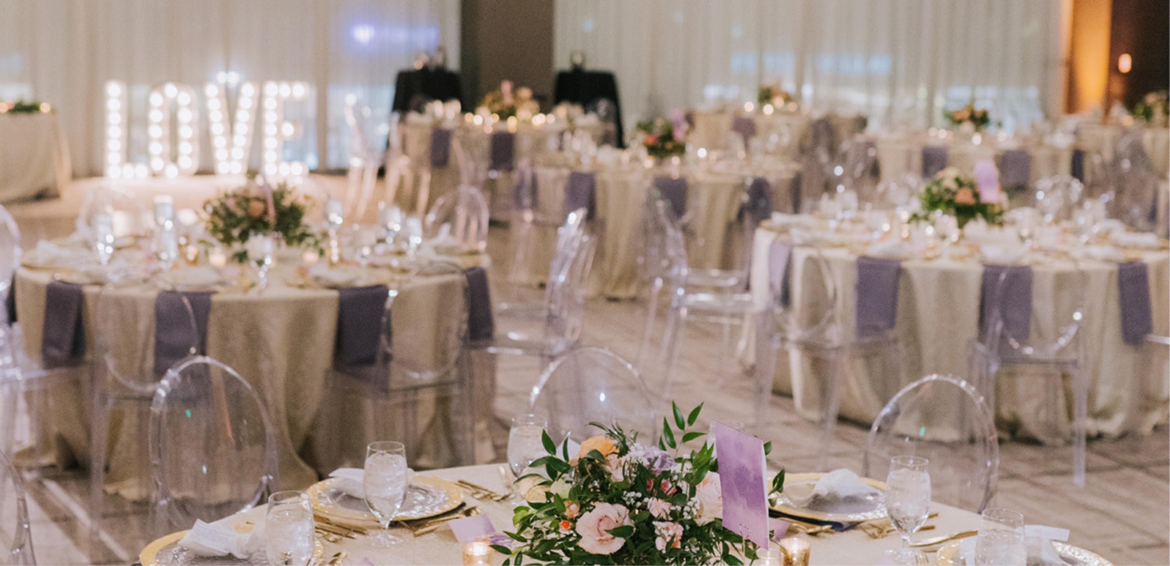 A ballroom with round tables dressed in gold and lilac linen, lilac napkins, candles and flowers, and a large lit-up 'LOVE' sign at the far end.