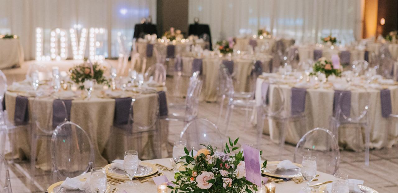 A ballroom with round tables dressed in gold and lilac linen, lilac napkins, candles and flowers, and a large lit-up 'LOVE' sign at the far end.