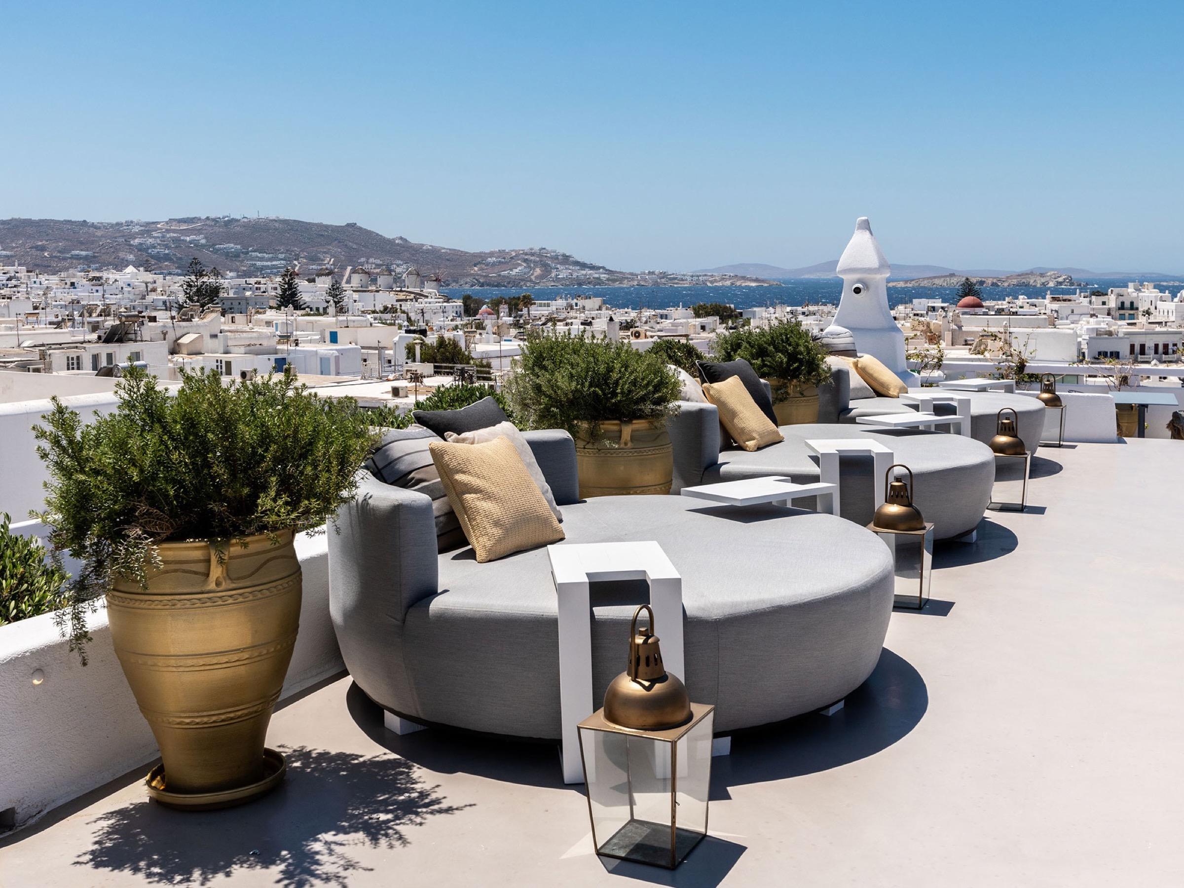 Round grey sofas and olive trees in brass pots on The TownHouse's roof, over the white roofs of Mykonos Town to the sea
