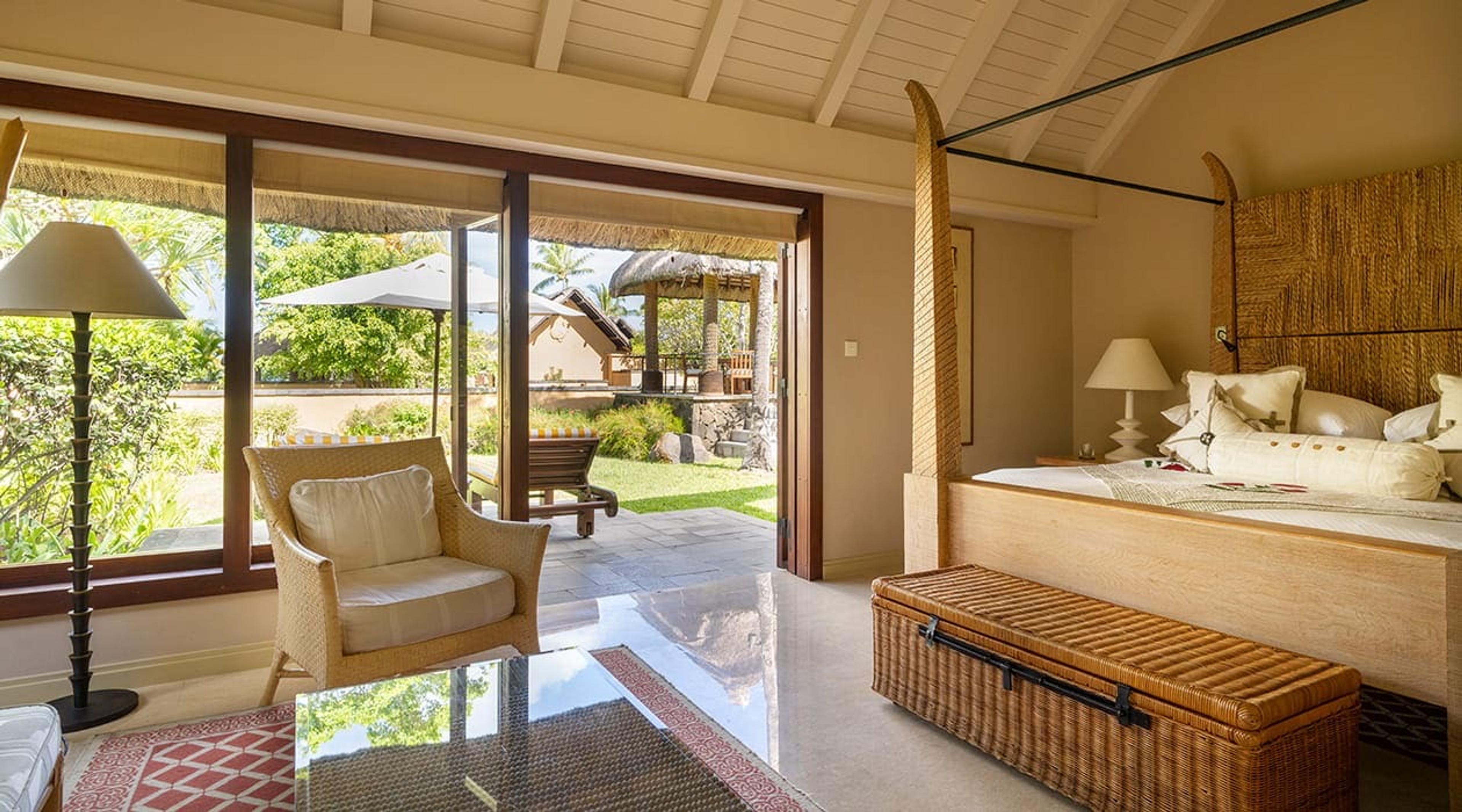 A bedroom with a four-poster bed and wicker armchair, with glass doors open onto a terrace, sun lounger and pool beyond.