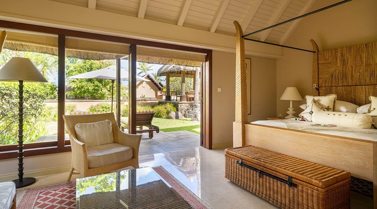 A bedroom with a four-poster bed and wicker armchair, with glass doors open onto a terrace, sun lounger and pool beyond.