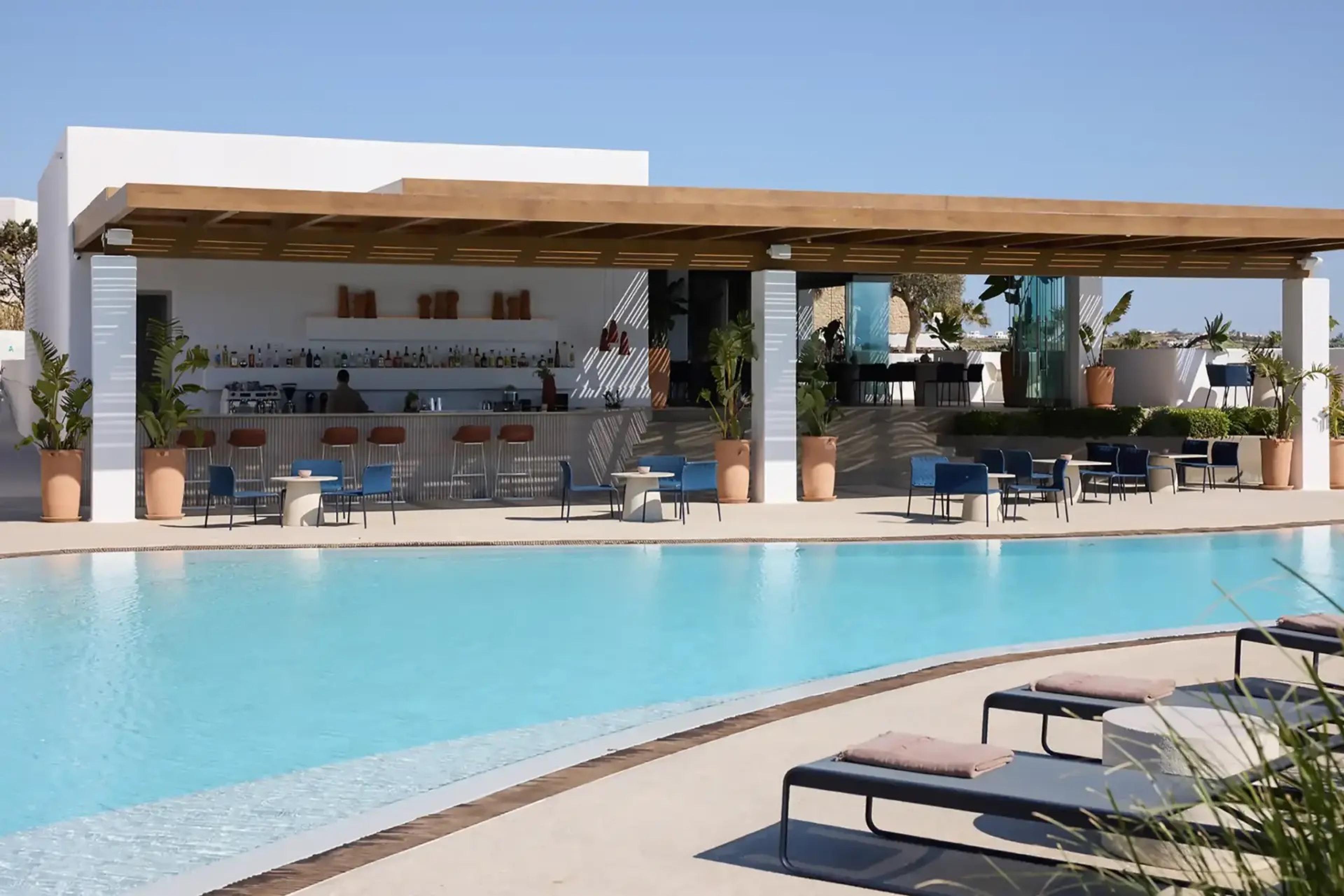 A poolside bar with a wooden pergola roof, shelves of bottles, blue outdoor chairs around tables, and sunbeds in the foreground under a clear sky.
