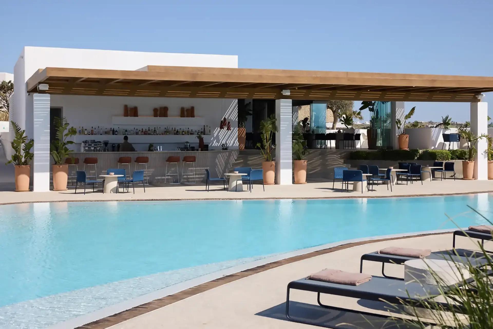 A poolside bar with a wooden pergola roof, shelves of bottles, blue outdoor chairs around tables, and sunbeds in the foreground under a clear sky.