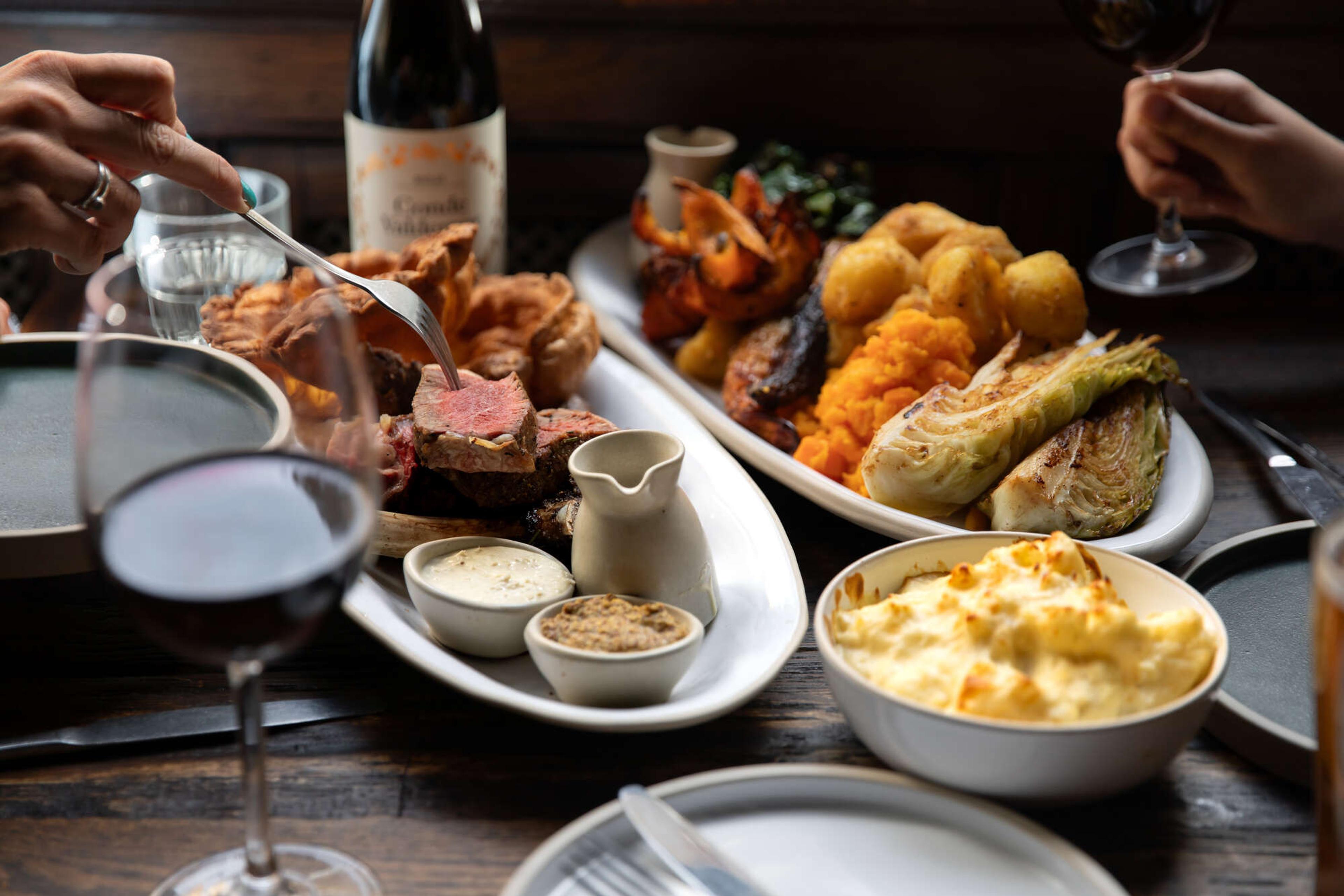 Two people reaching across a table laid with a Sunday roast, including a sliced joint of beef, roast vegetables, Yorkshire puddings and a bowl of cauliflower cheese.