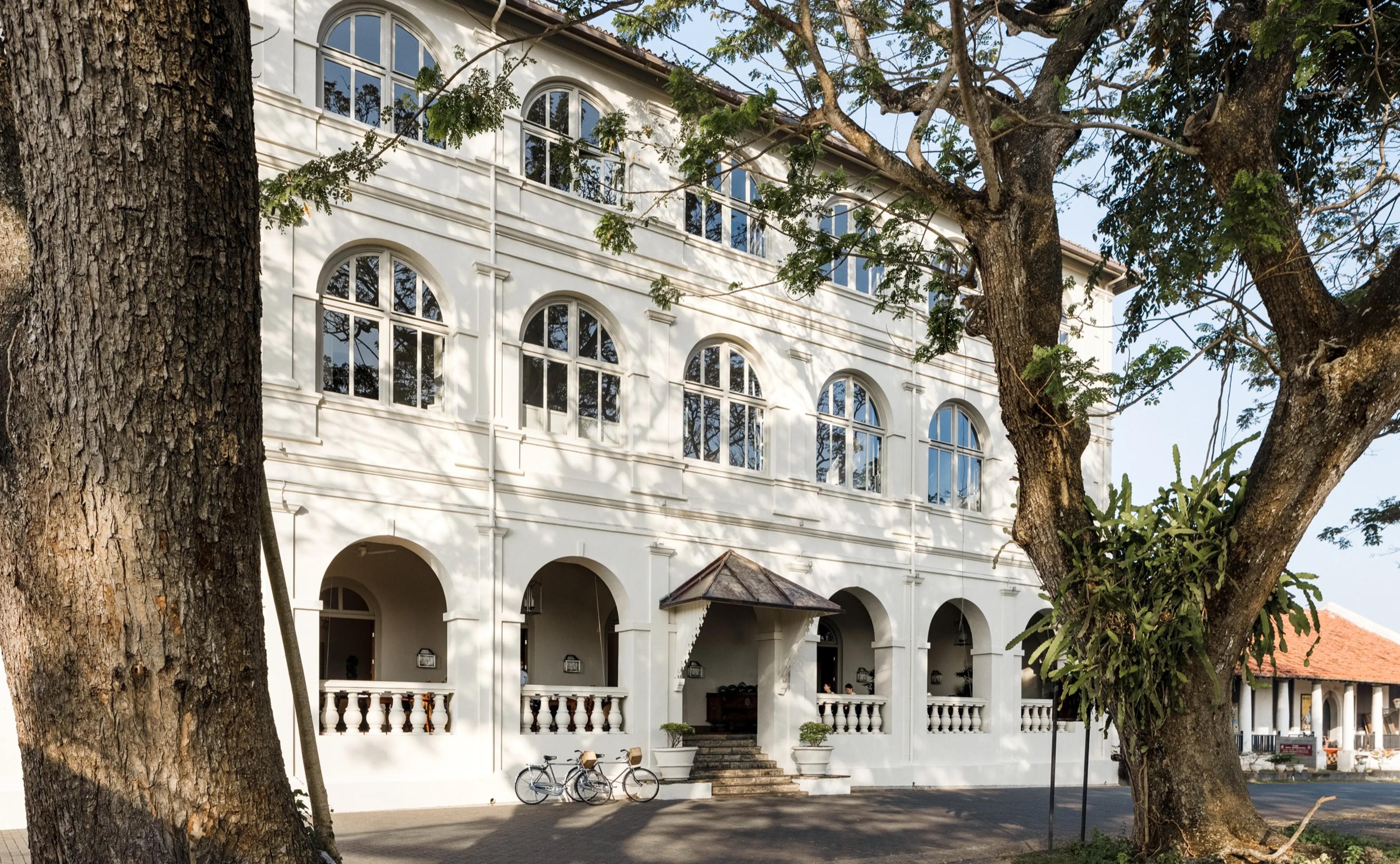 A three-storey colonial building with arched windows and a columned entrance porch, framed by two large trees with bicycles parked at the foot of the steps.