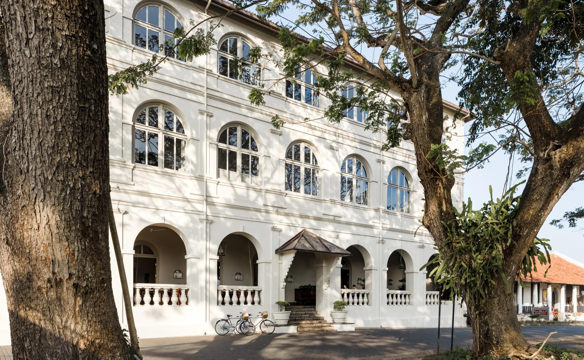 A three-storey colonial building with arched windows and a columned entrance porch, framed by two large trees with bicycles parked at the foot of the steps.