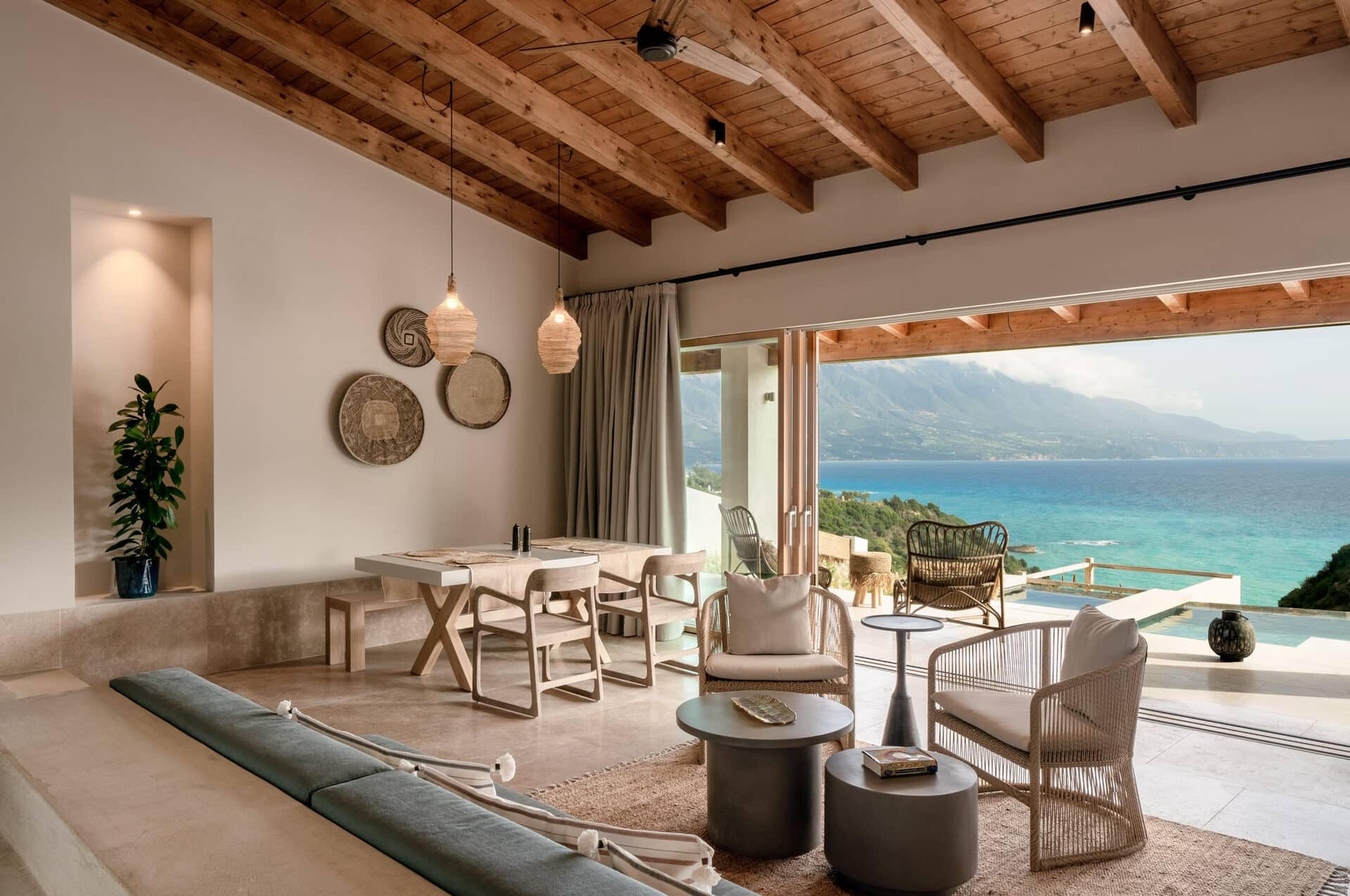 an open plan living and dining room with a timber beamed ceiling, a dining table, woven wall plates and armchairs, opening onto a terrace with a sea view