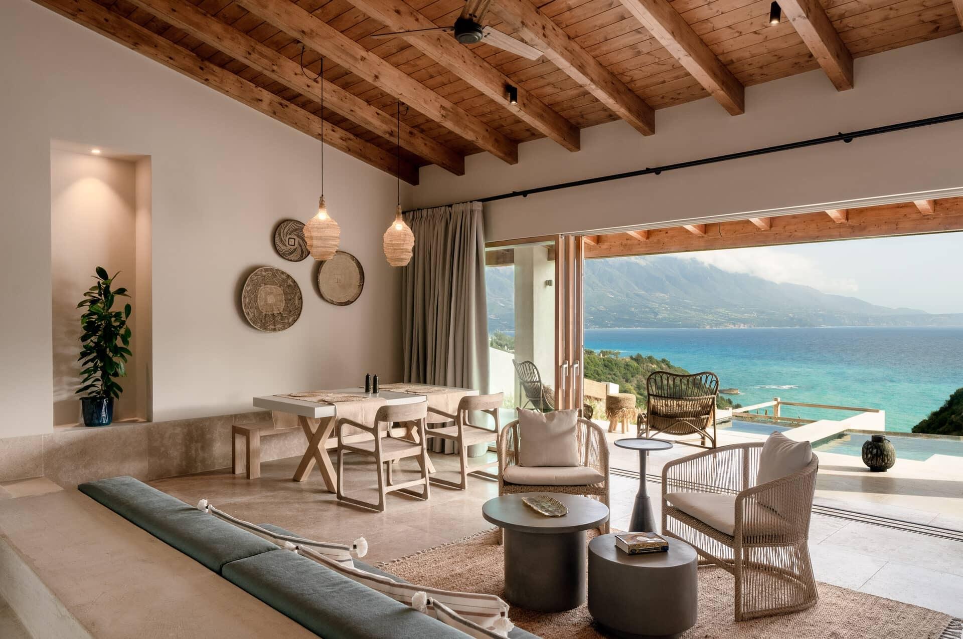 an open plan living and dining room with a timber beamed ceiling, a dining table, woven wall plates and armchairs, opening onto a terrace with a sea view