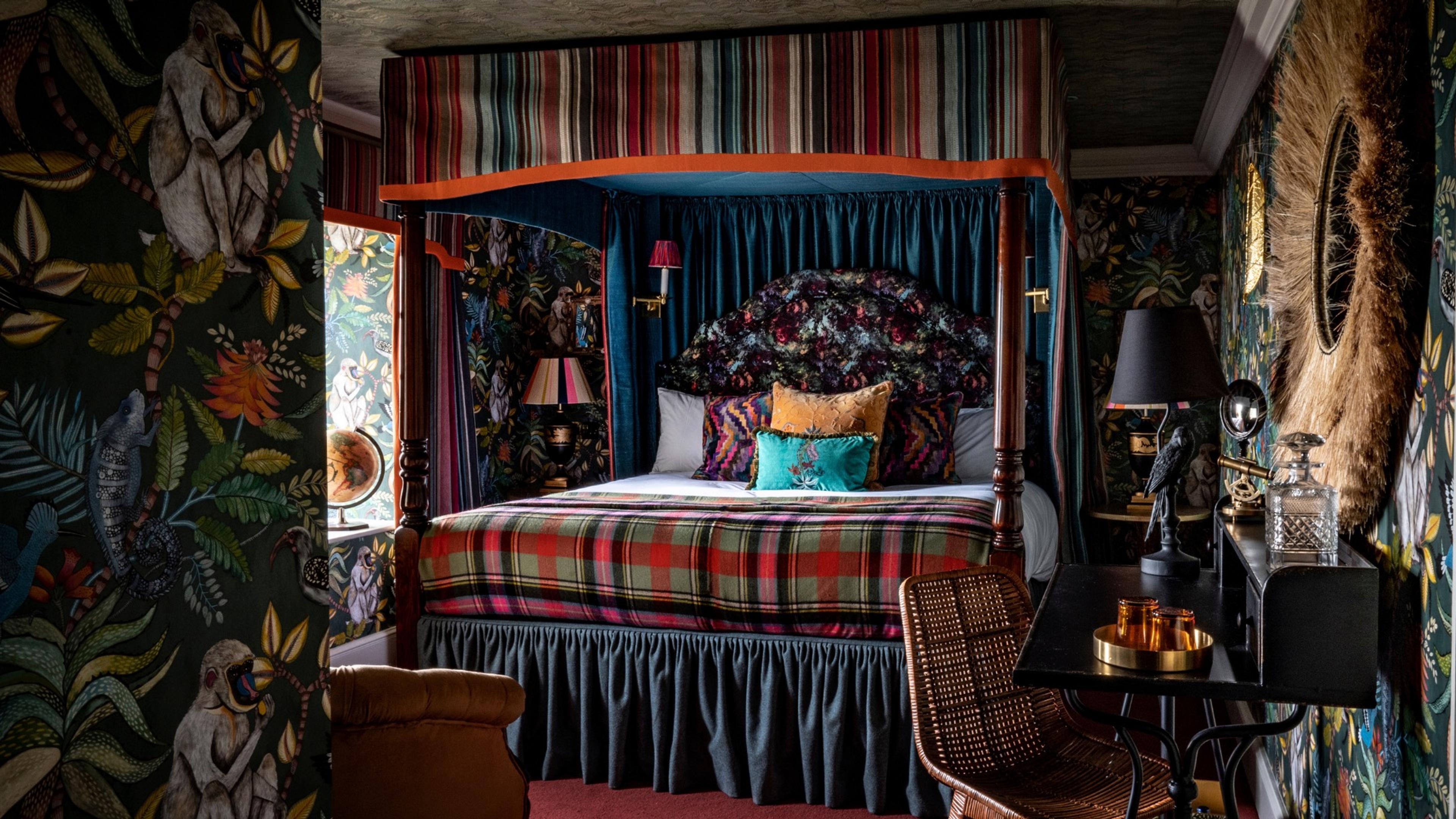 A four poster bed with a striped canopy and tartan blanket in a bedroom with dark jungle-print wallpaper and a wicker chair