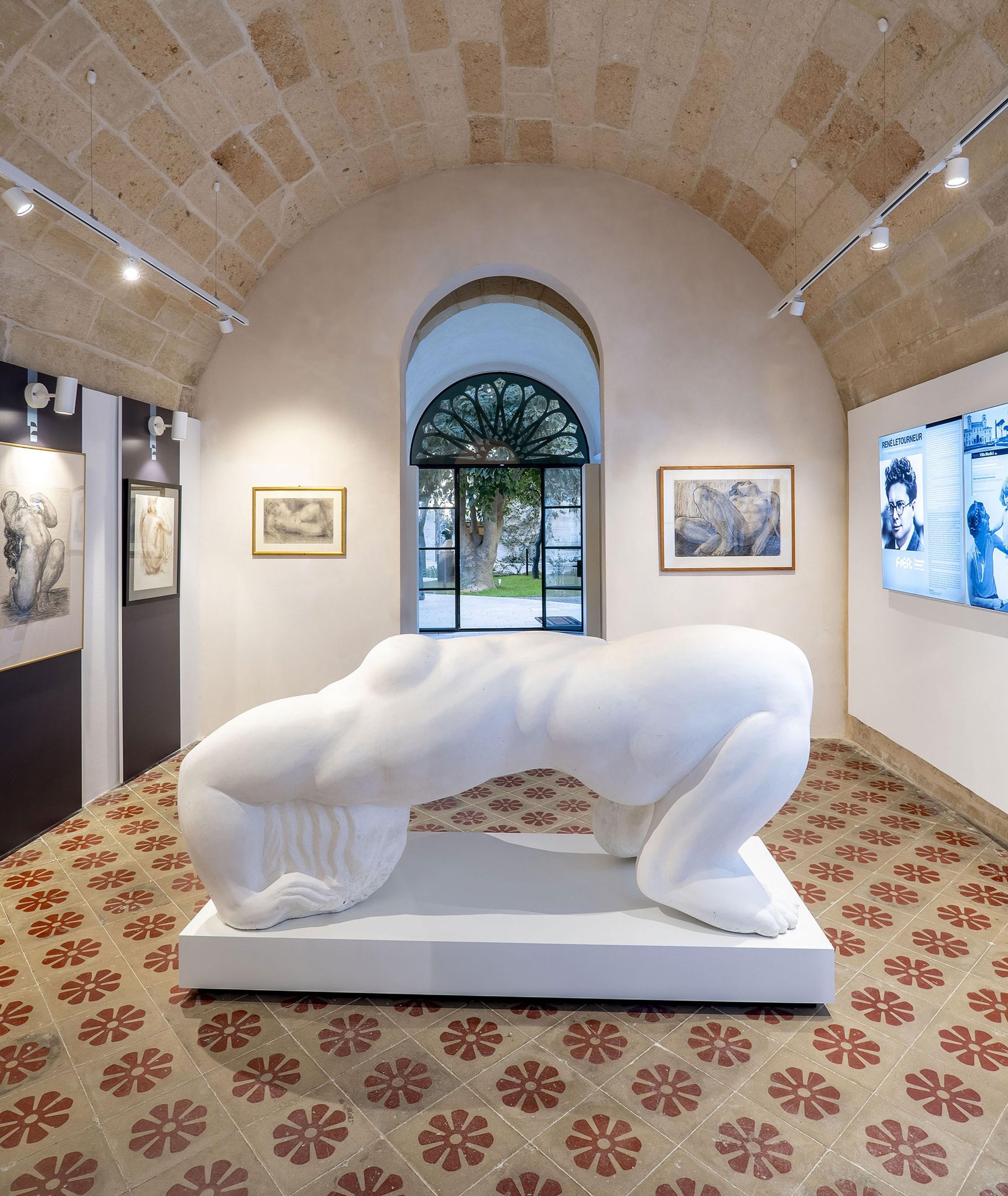 A vaulted gallery room with a large reclining nude sculpture on a white plinth, framed drawings on the walls, and an arched window onto a garden.