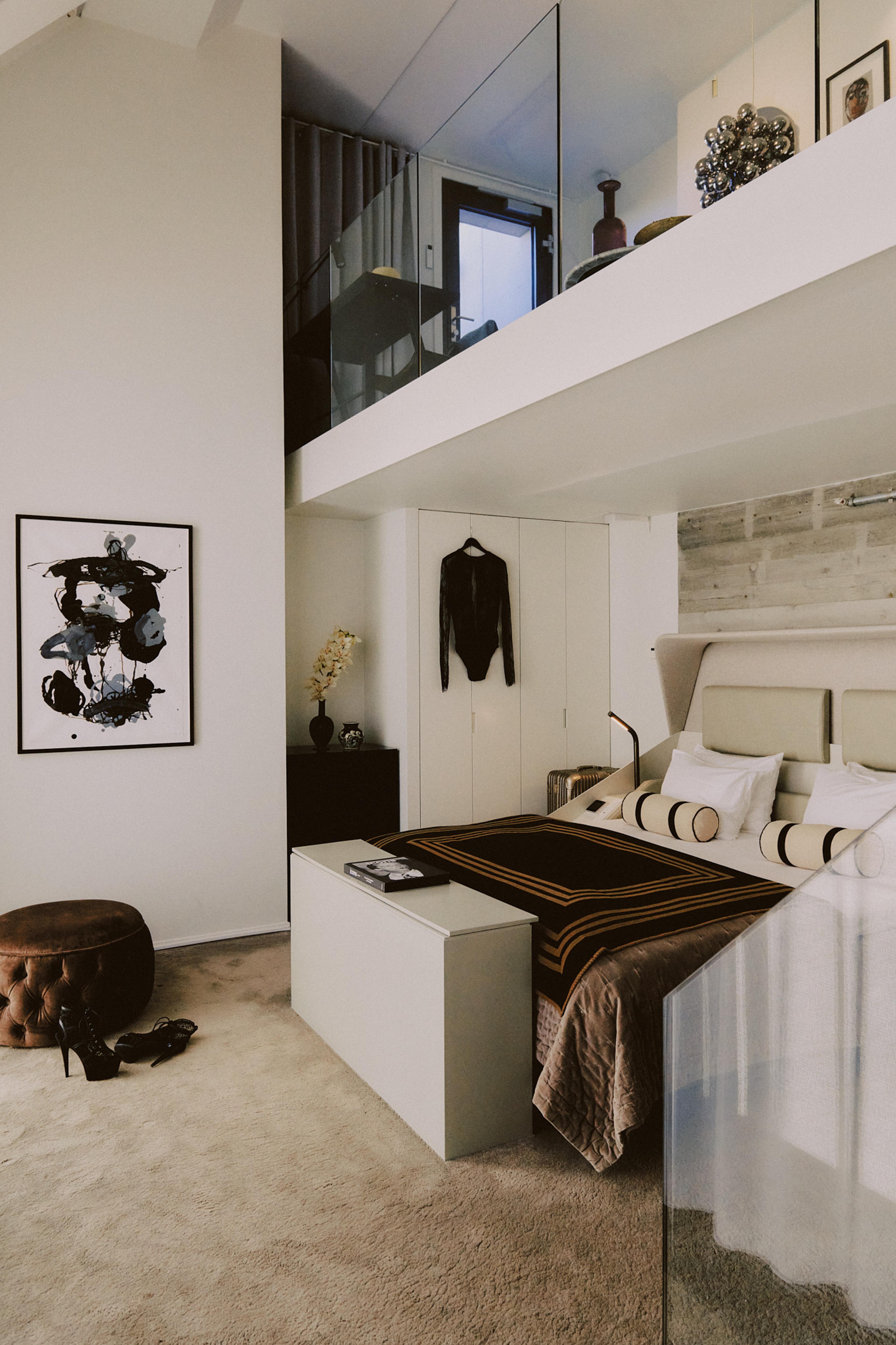 A bedroom with a mezzanine above, a bed with a patterned brown throw, an abstract painting on the wall, a pair of boots and heels on the floor, and a jacket on a hanger.
