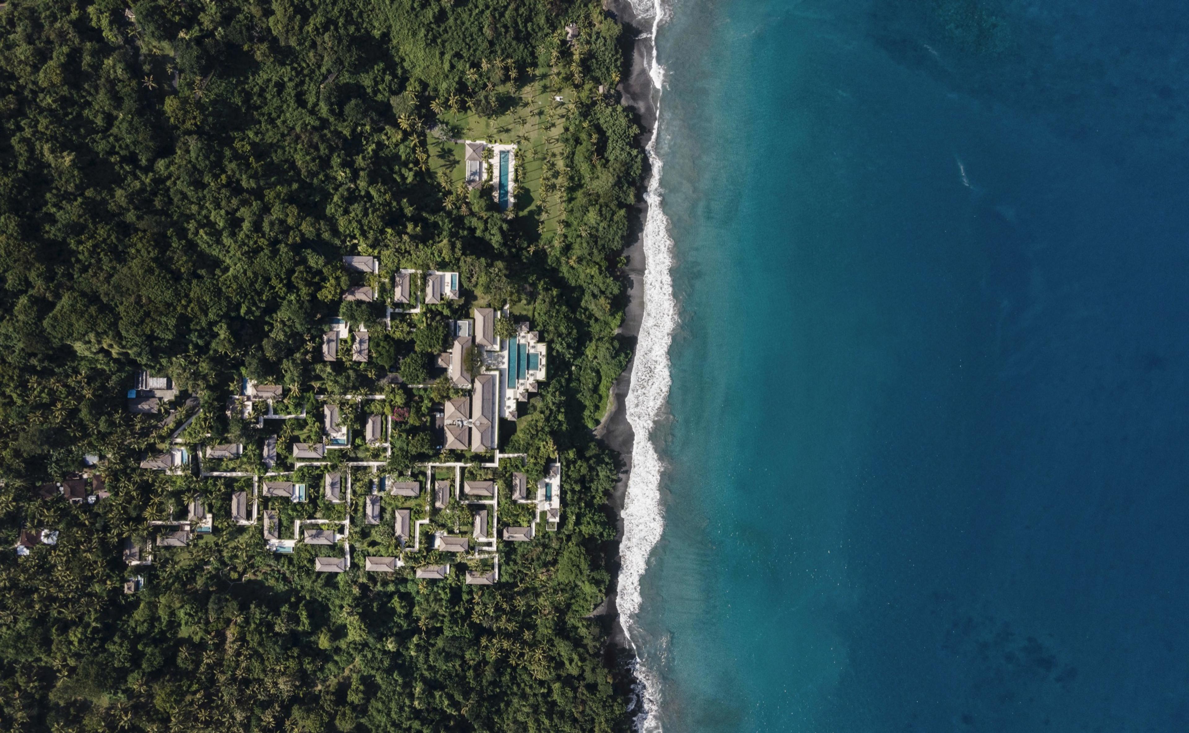 An aerial view of a hillside resort of thatched roofs and pools set among dense jungle along a black sand coastline.