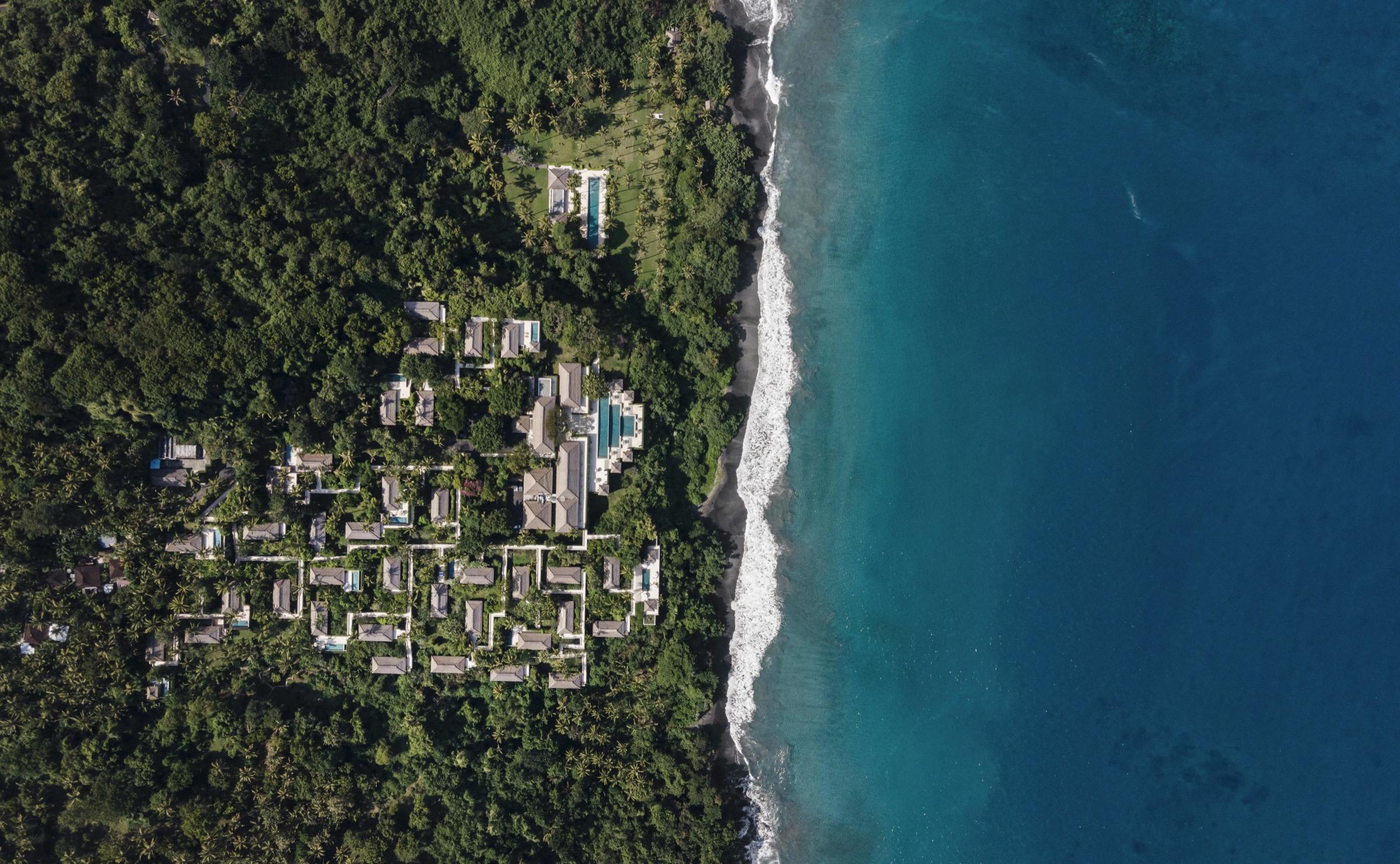 An aerial view of a hillside resort of thatched roofs and pools set among dense jungle along a black sand coastline.