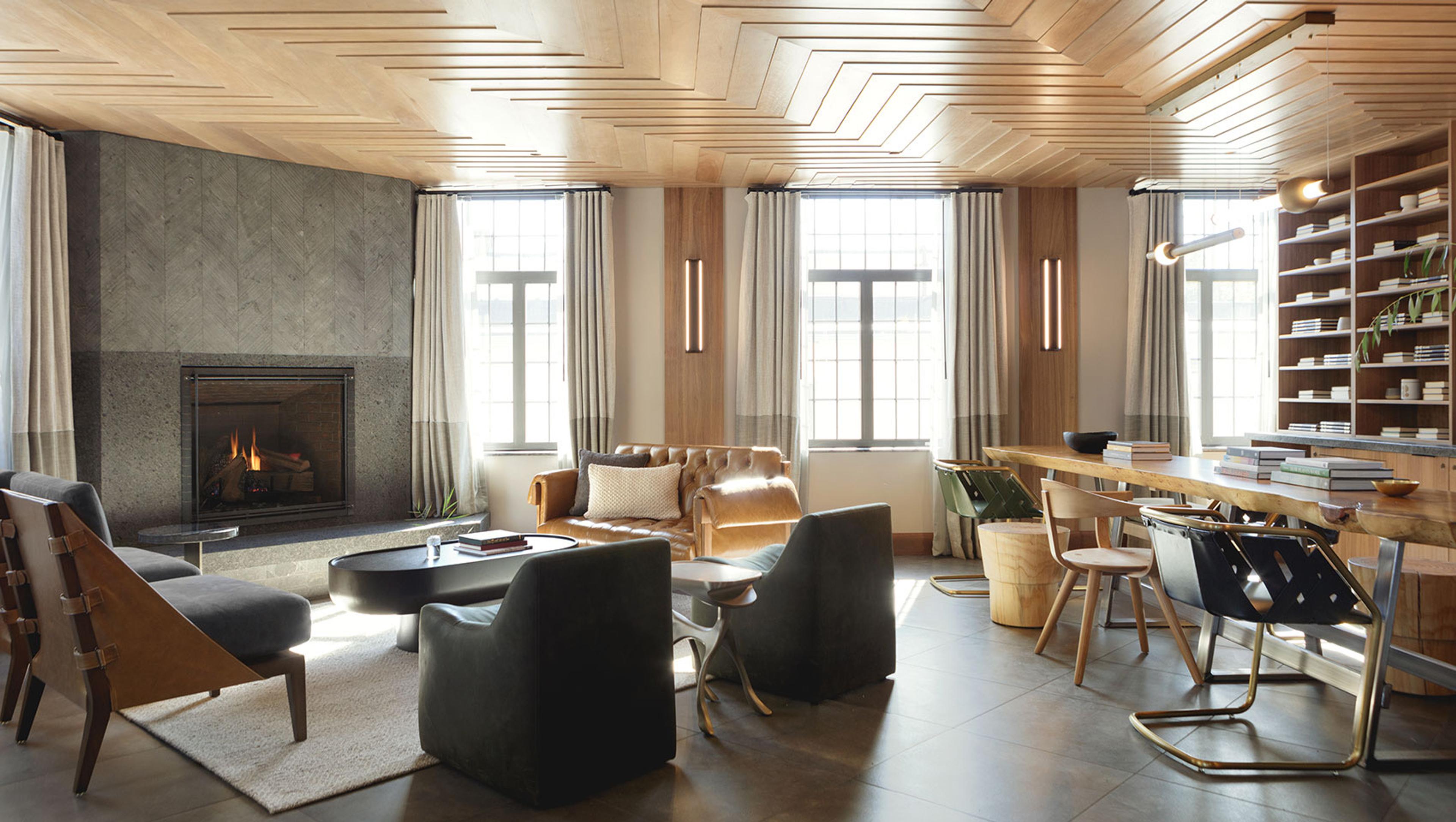 A hotel lounge with a stone fireplace, leather and canvas armchairs on a rug, and a long communal table in front of a wall of shelved books.