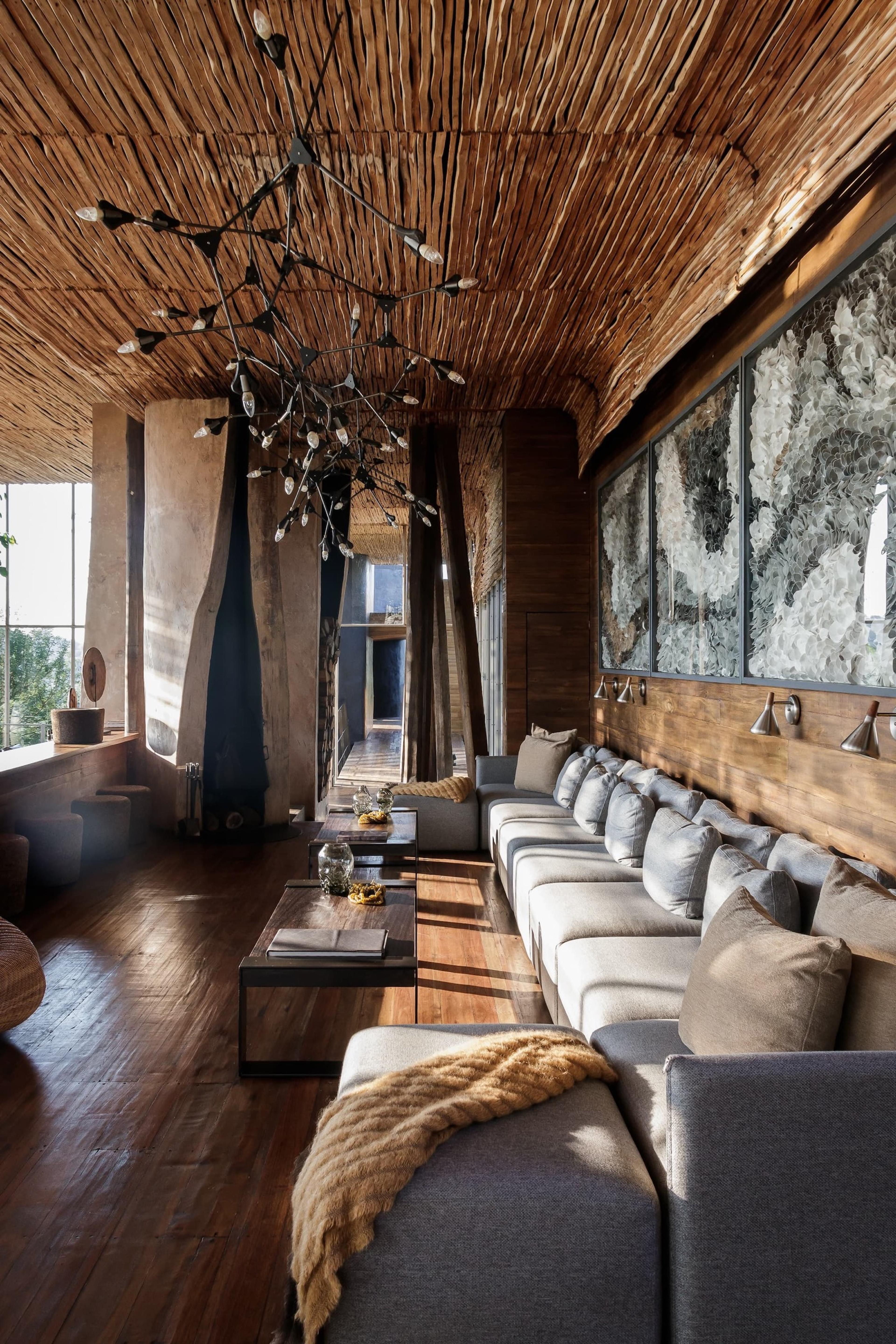 A long grey sectional sofa sits beneath a reed ceiling and a branch-like black chandelier, facing floor-to-ceiling windows onto greenery.
