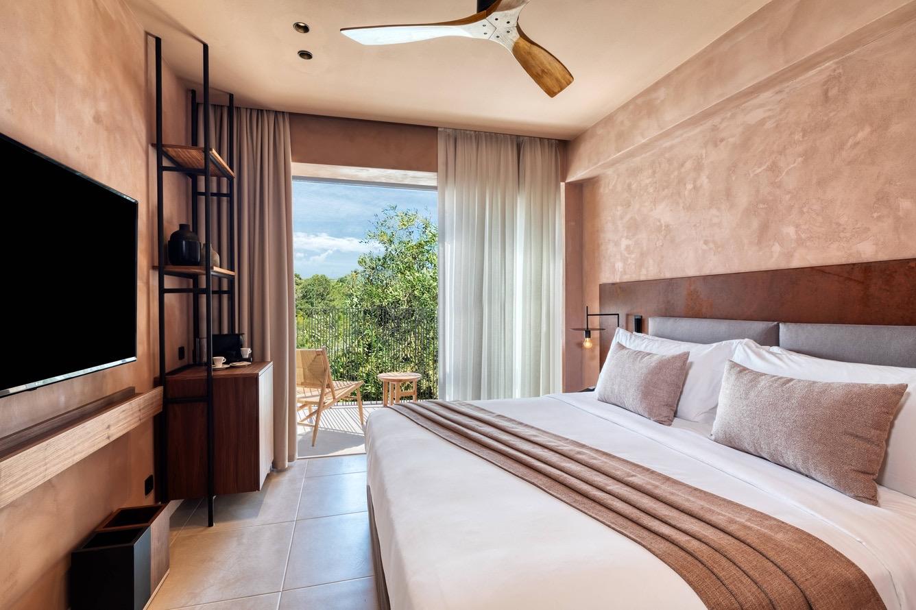 A hotel bedroom with a wall-mounted television, open shelving, and a bed with brown pillows, facing a balcony open to trees.