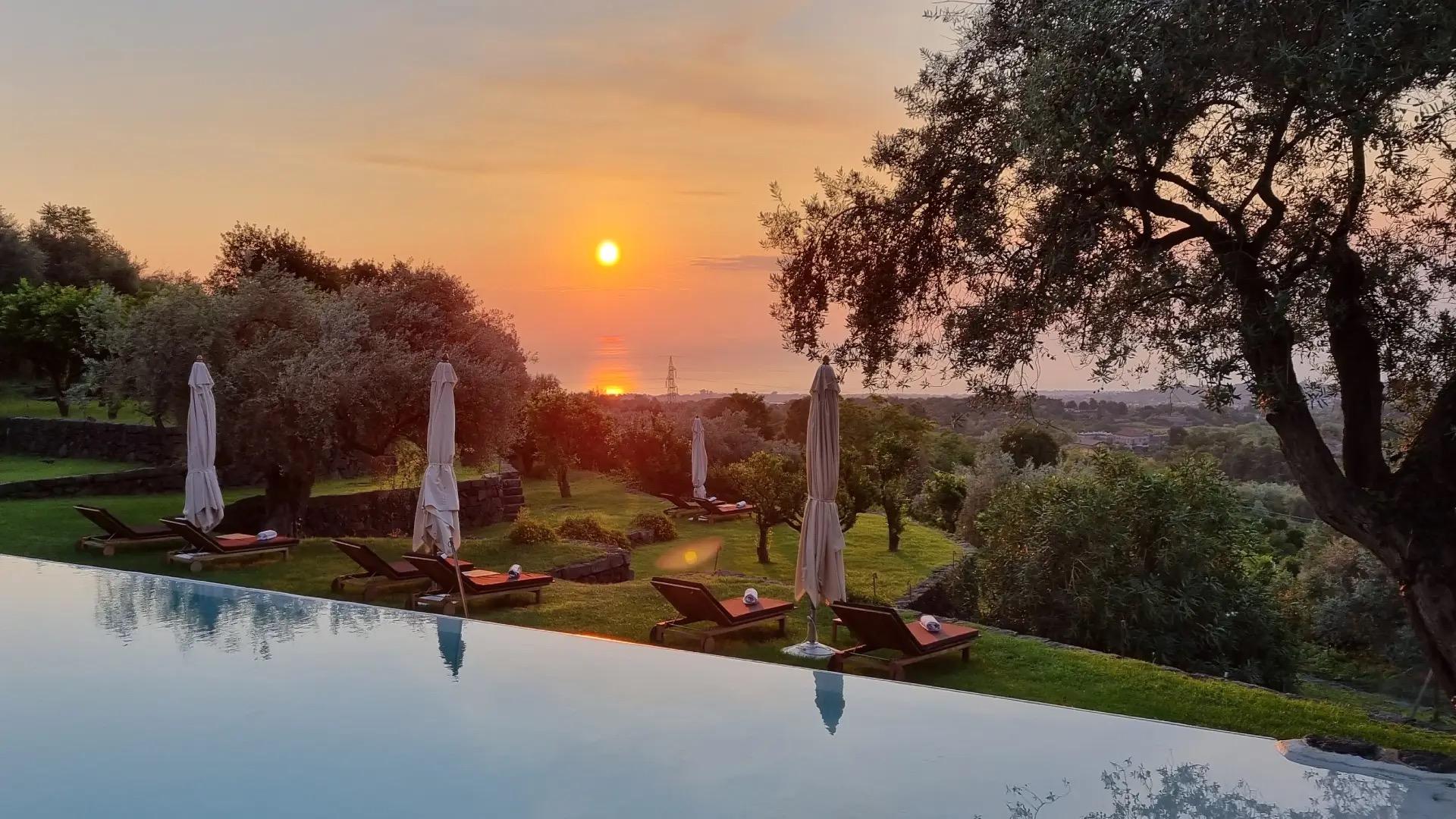 Sun loungers and furled umbrellas beside an infinity pool at sunset, with the sun low over the sea.