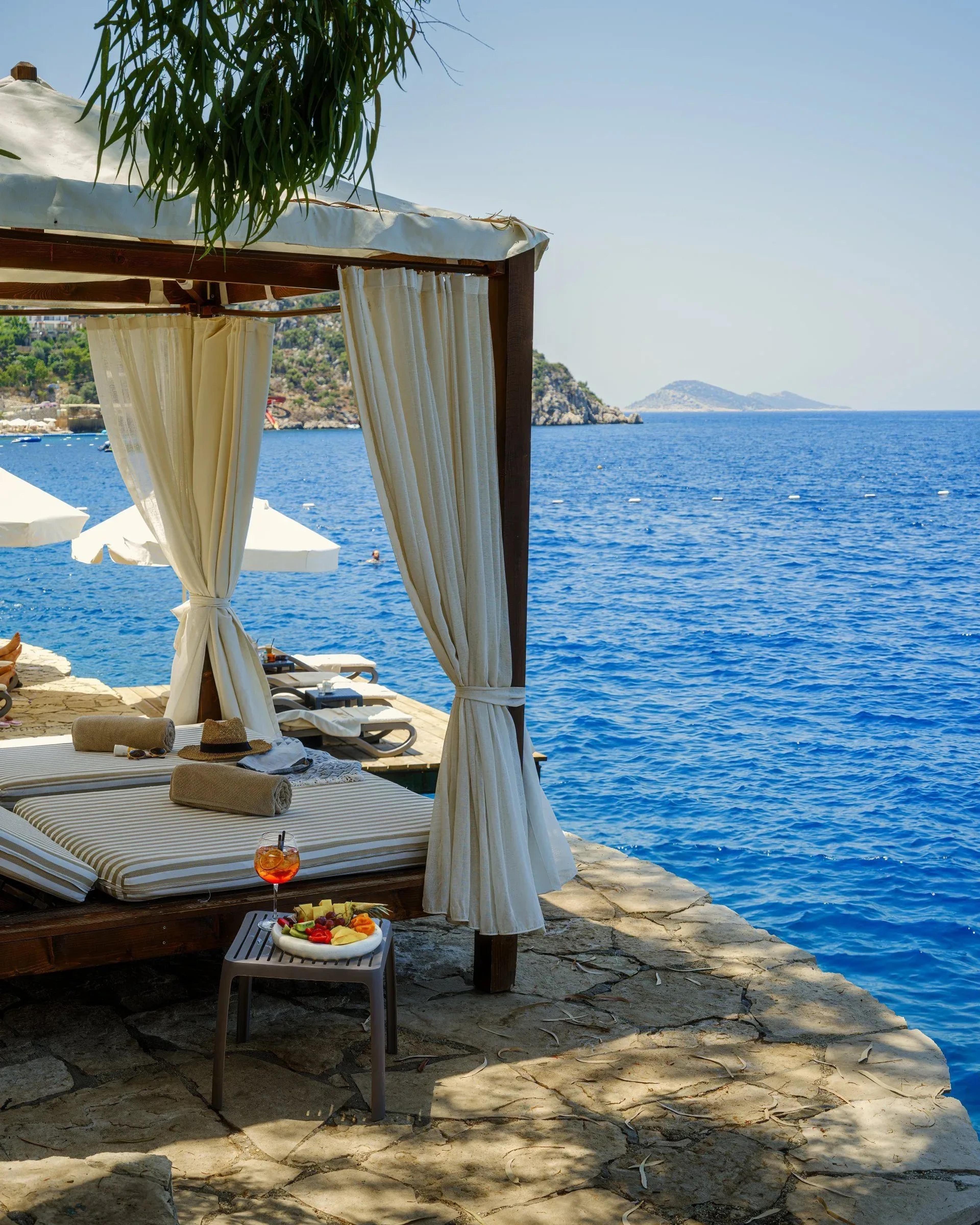 A cabana with white curtains and daybeds on a stone terrace at the water's edge, with a fruit platter and an orange spritz on a low table and an island beyond.
