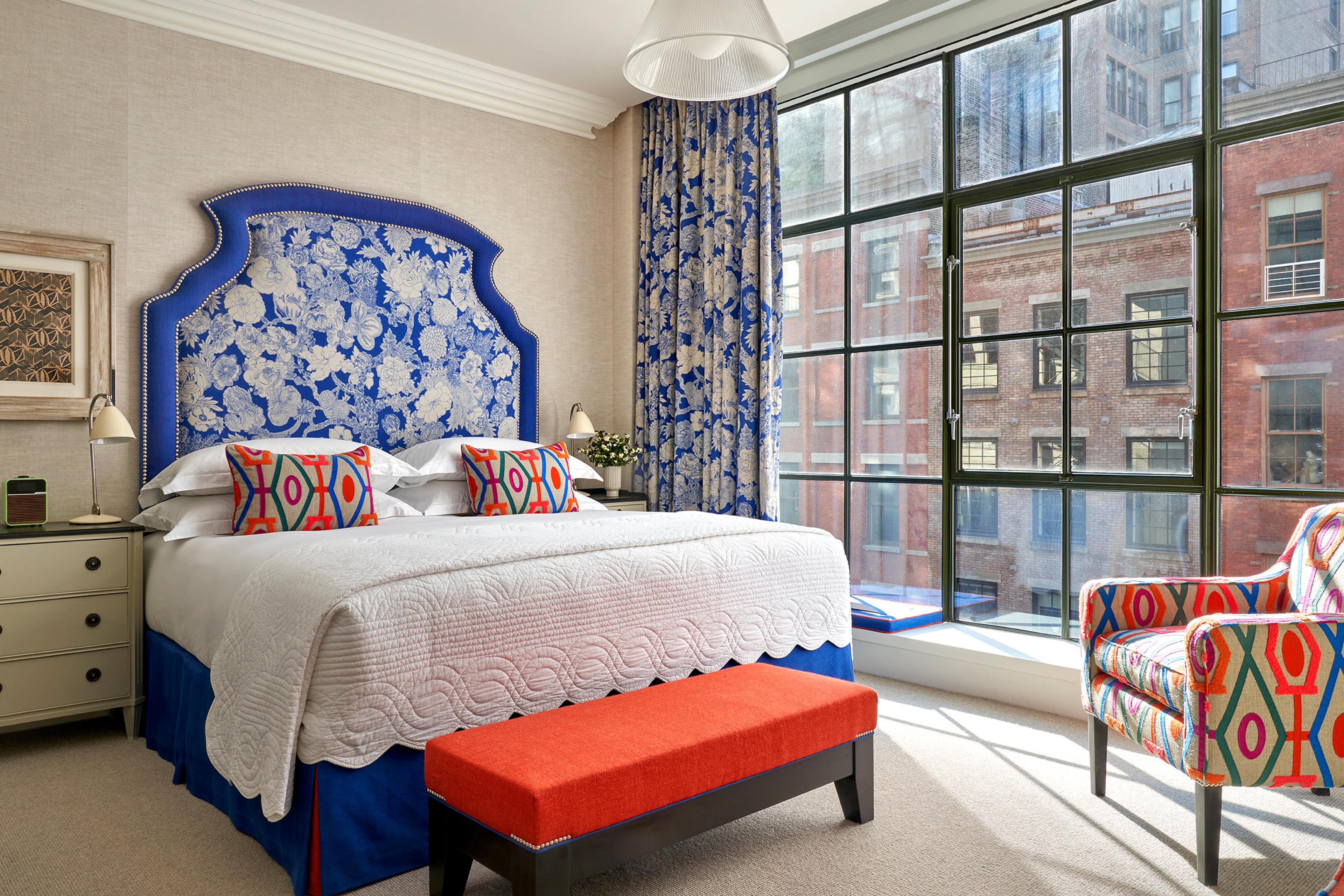 A bedroom with a blue floral headboard, an orange bench at the foot of the bed, and floor-to-ceiling windows looking onto brick buildings across the street.