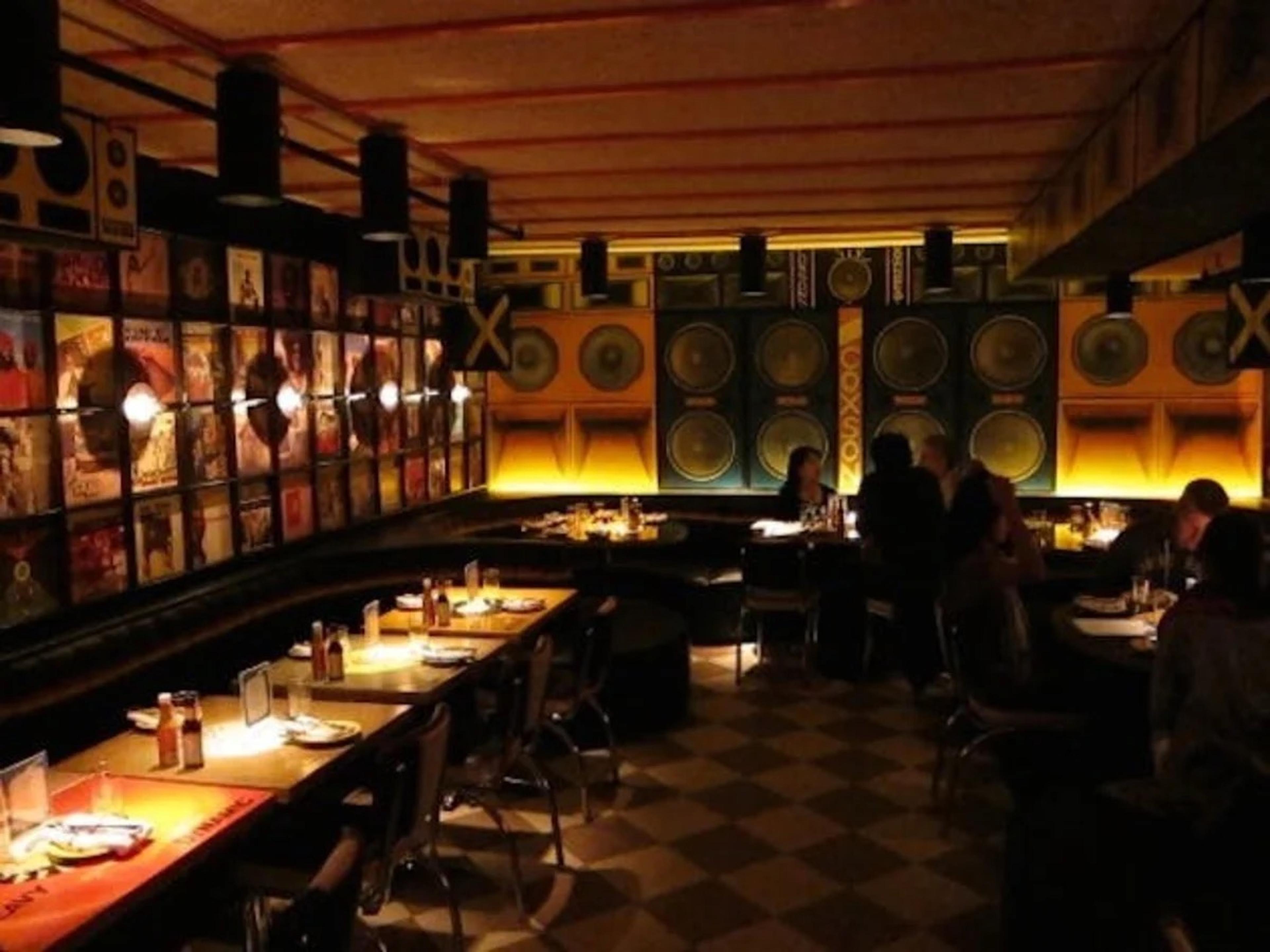 A dim dining room with a long banquette, warm lamps and record sleeves lining the wall