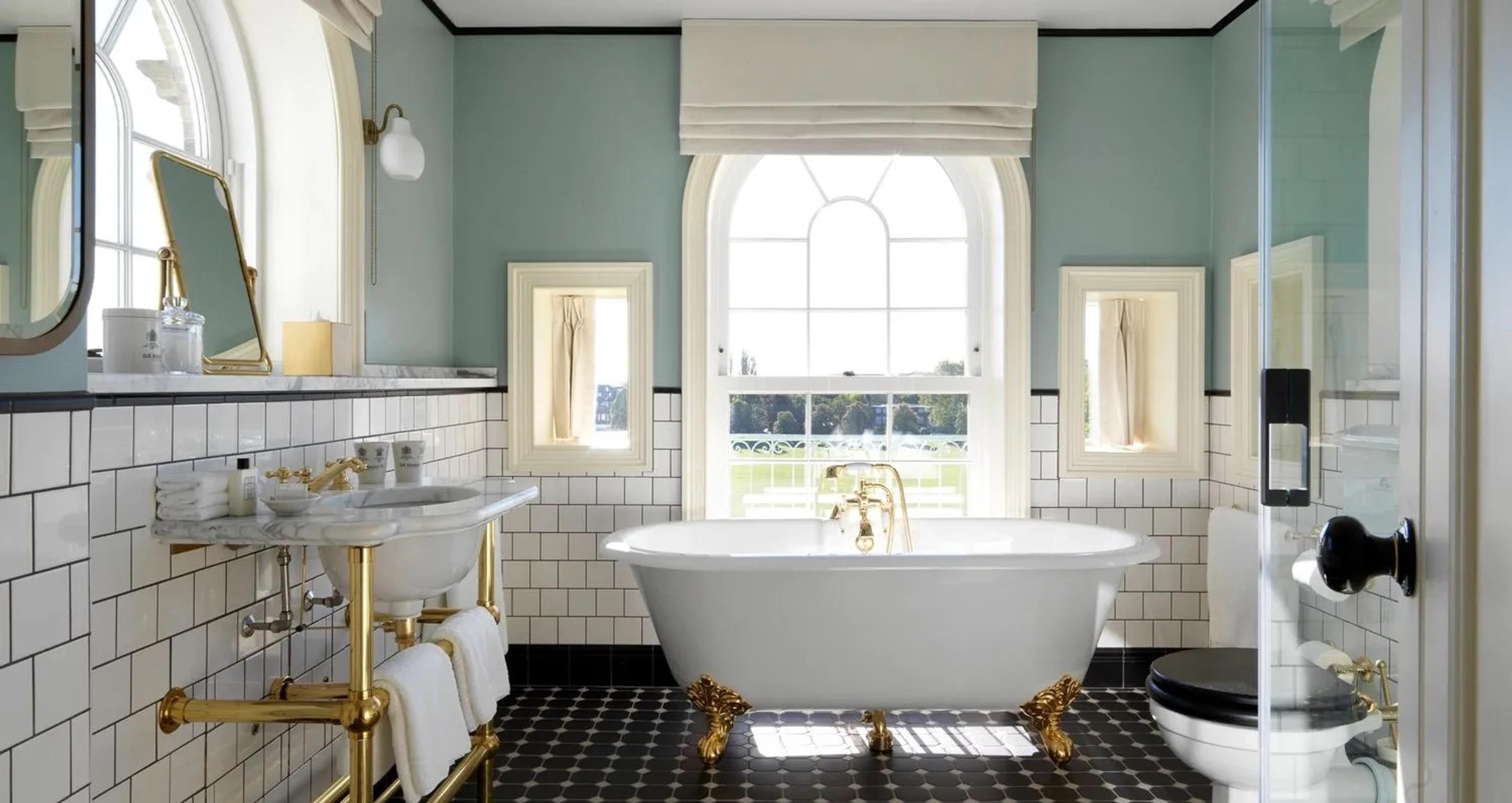 a bathroom with white metro tiles, a black and white tiled floor, a freestanding white bathtub on gold feet, and an arched window flanked by two smaller square windows