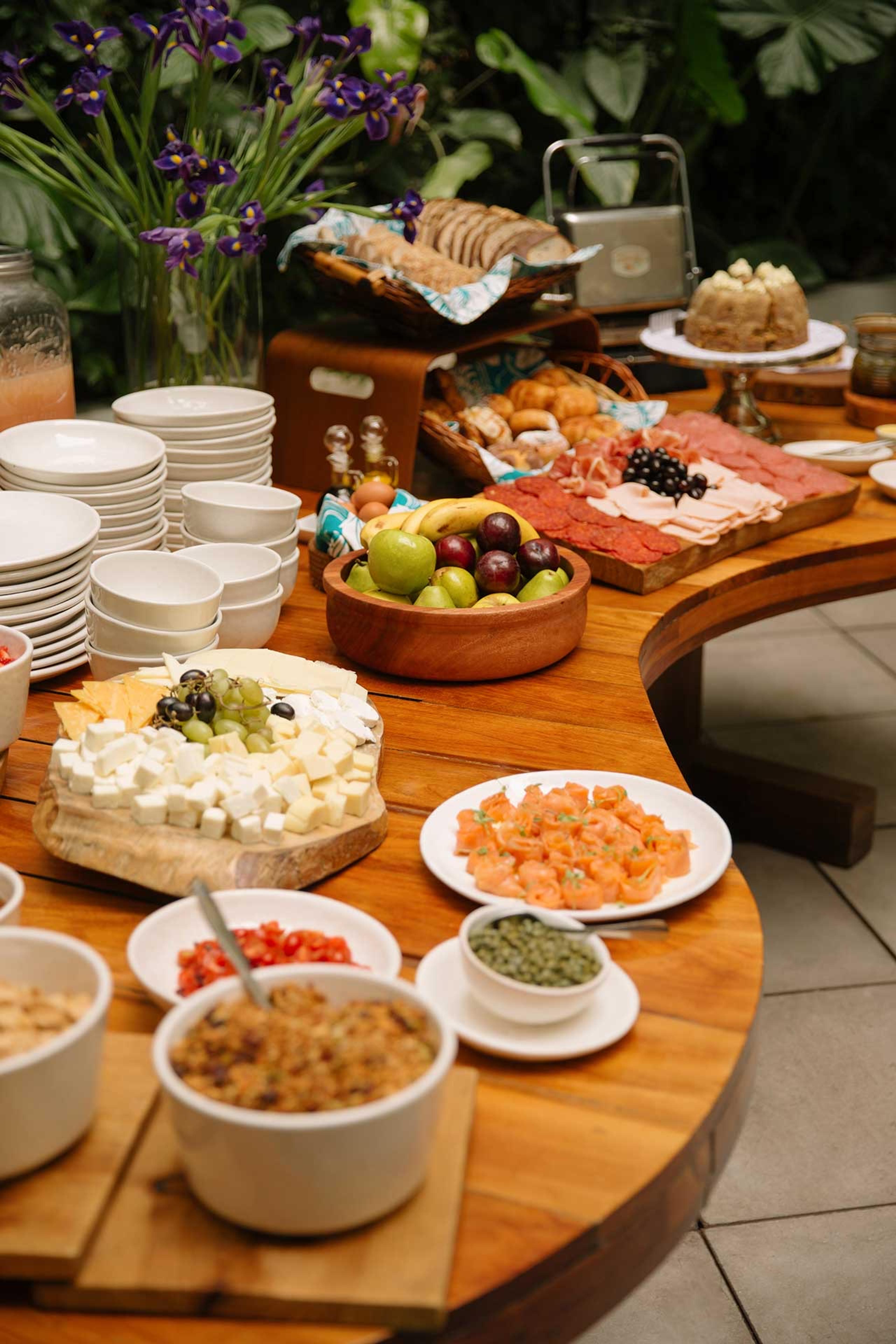 A wooden table spread with cheese, charcuterie, fruit, pastries, bread and small bowls of condiments, with purple flowers in the background.