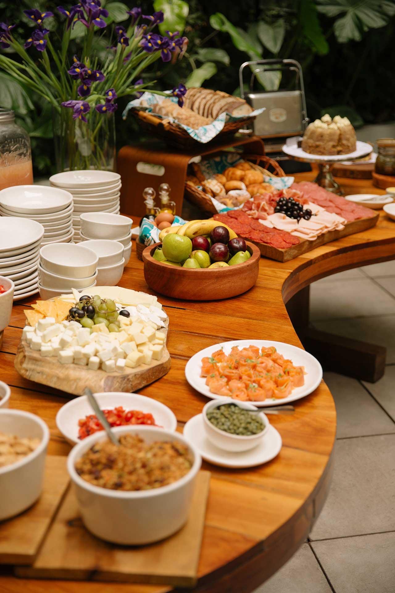 A wooden table spread with cheese, charcuterie, fruit, pastries, bread and small bowls of condiments, with purple flowers in the background.