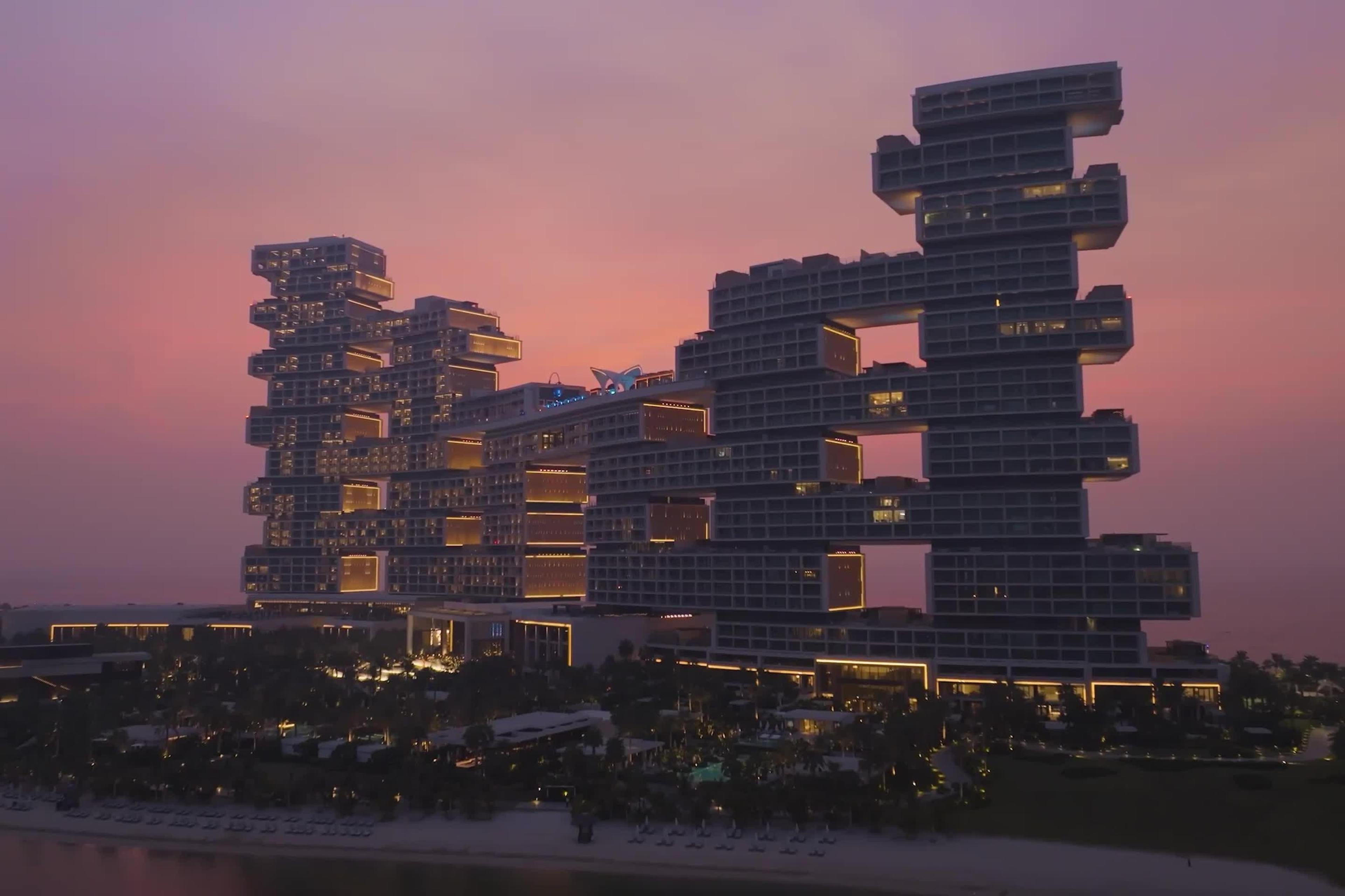The hotel's two arched towers rise above a lit beach and pool garden at dusk, seen from the air.