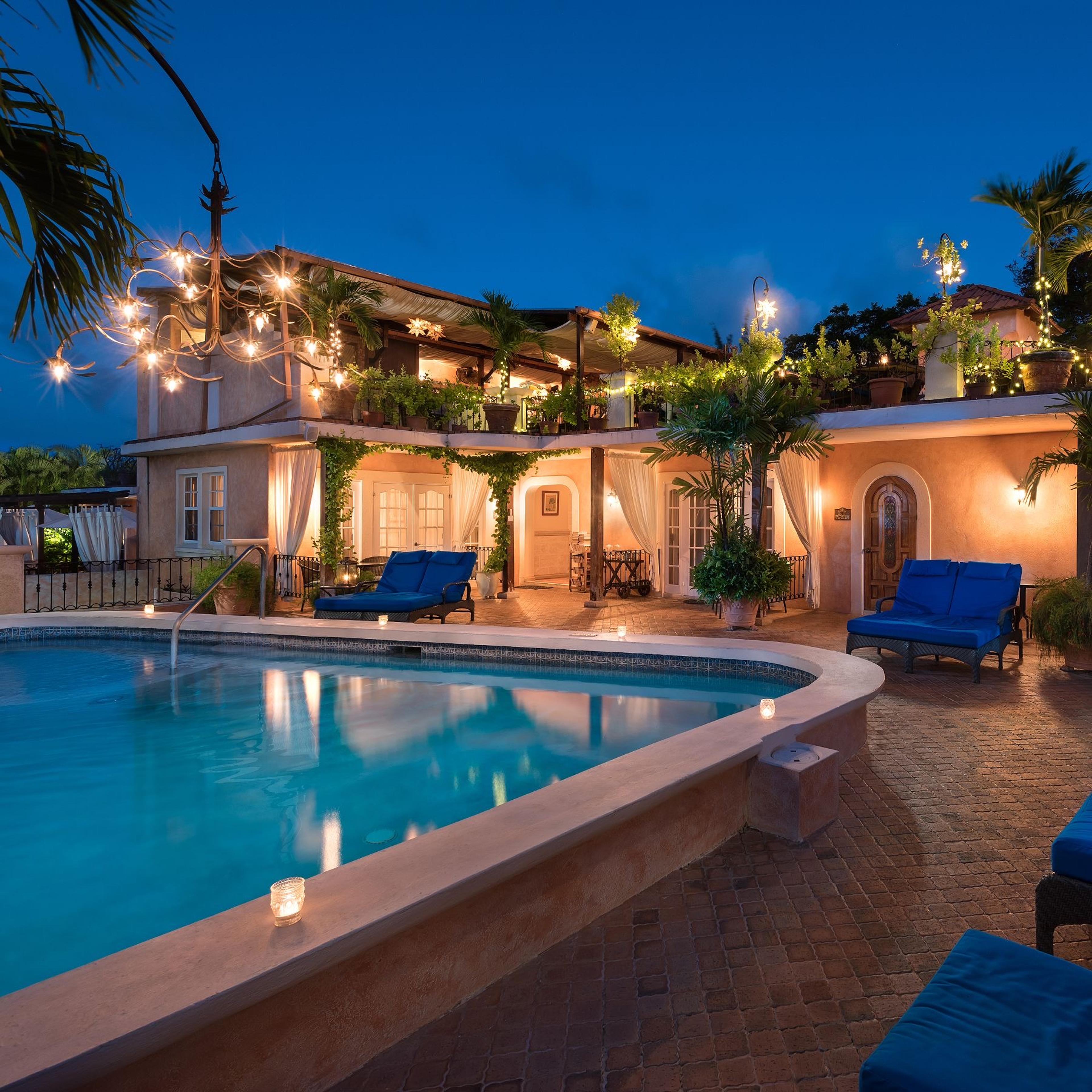 The hotel building and swimming pool at dusk, lit by string lights and candles along the pool edge, with blue loungers on the terrace.