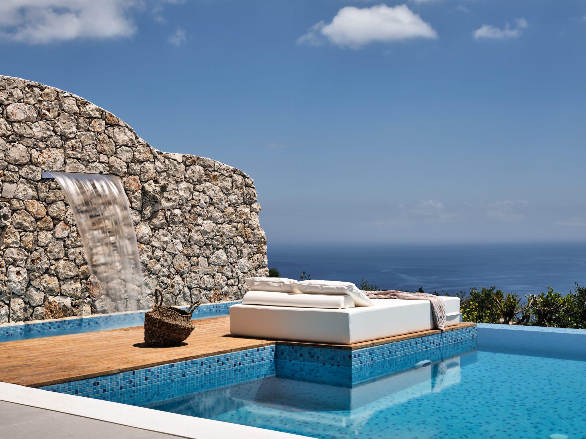 a built-in daybed on a pool's edge beside a curved stone wall with water running down it, the sea beyond