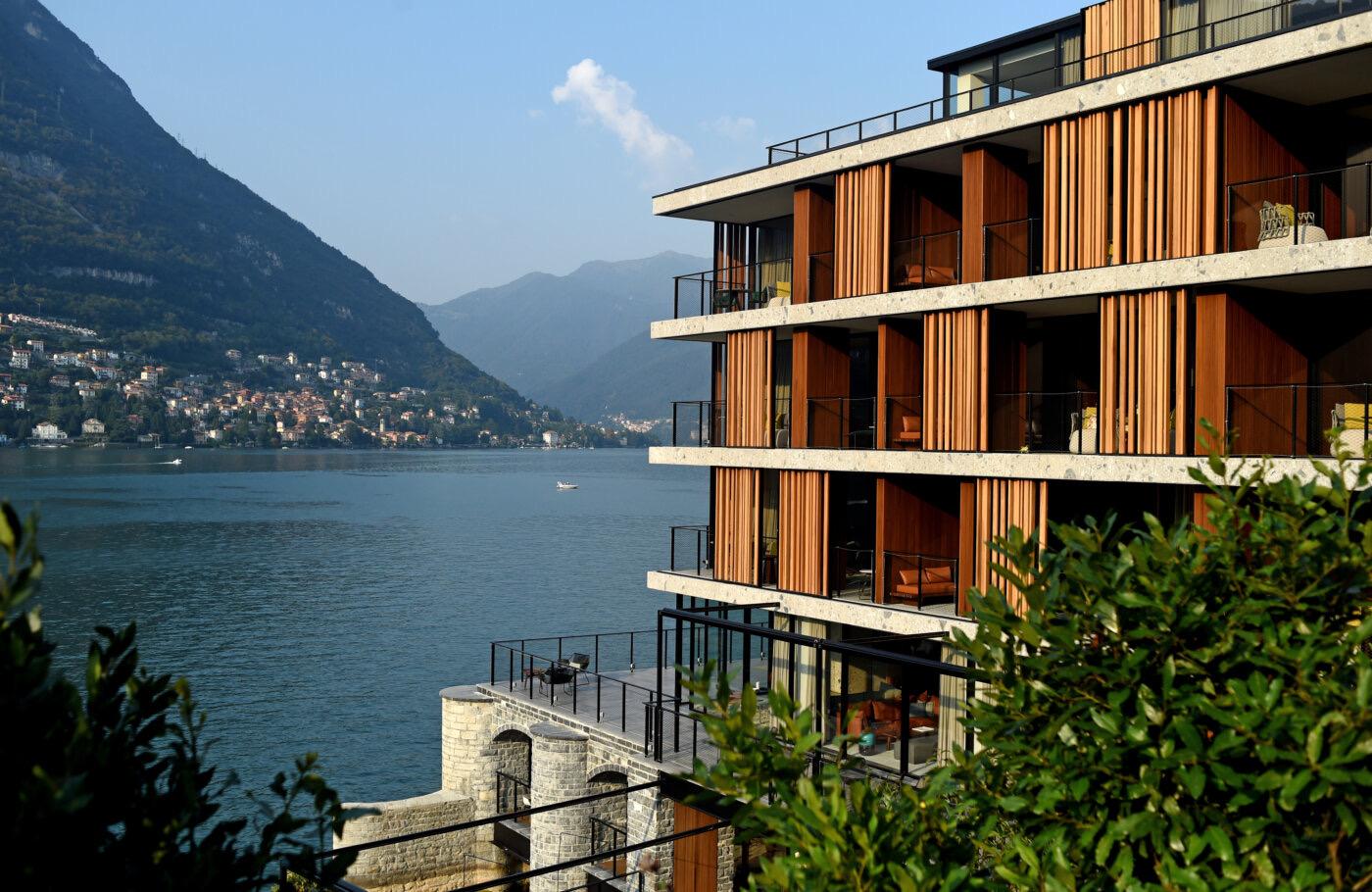 The corner of a hotel building seen from across the water, its floors wrapped in timber slat balconies above a stone base, with trees in the foreground.