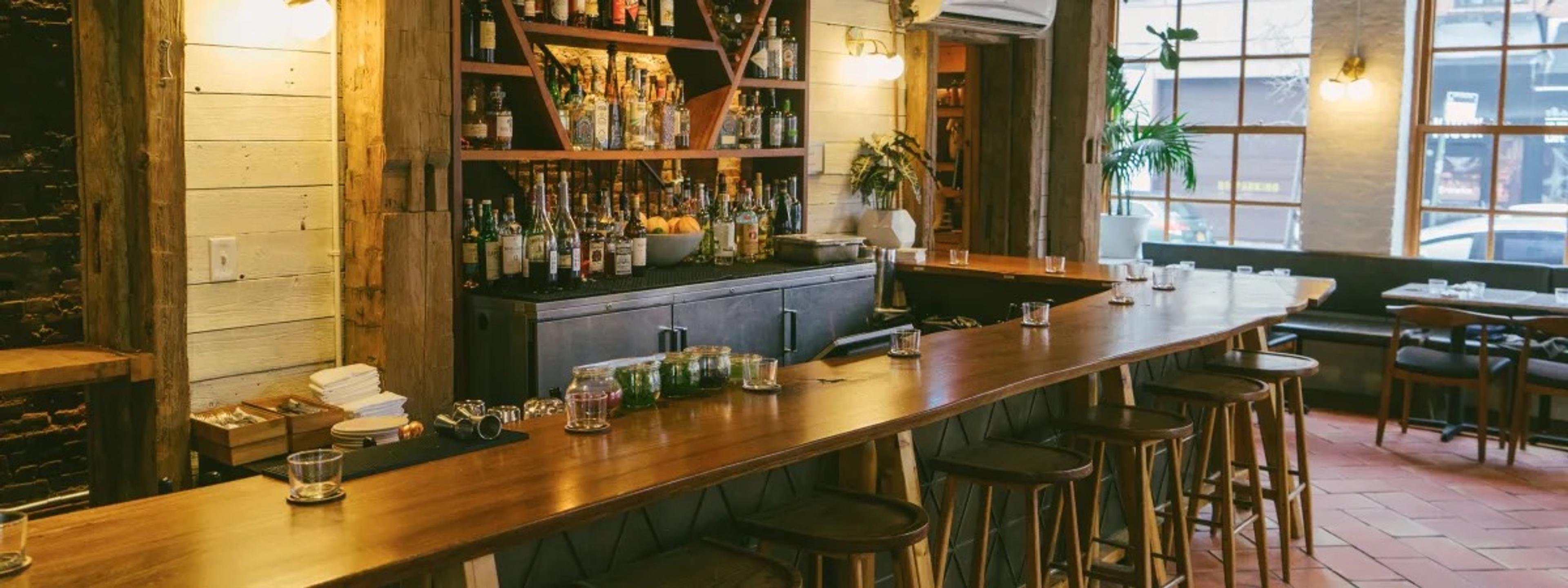 A long wooden bar with stools, shelves of bottles behind it and plants in the window