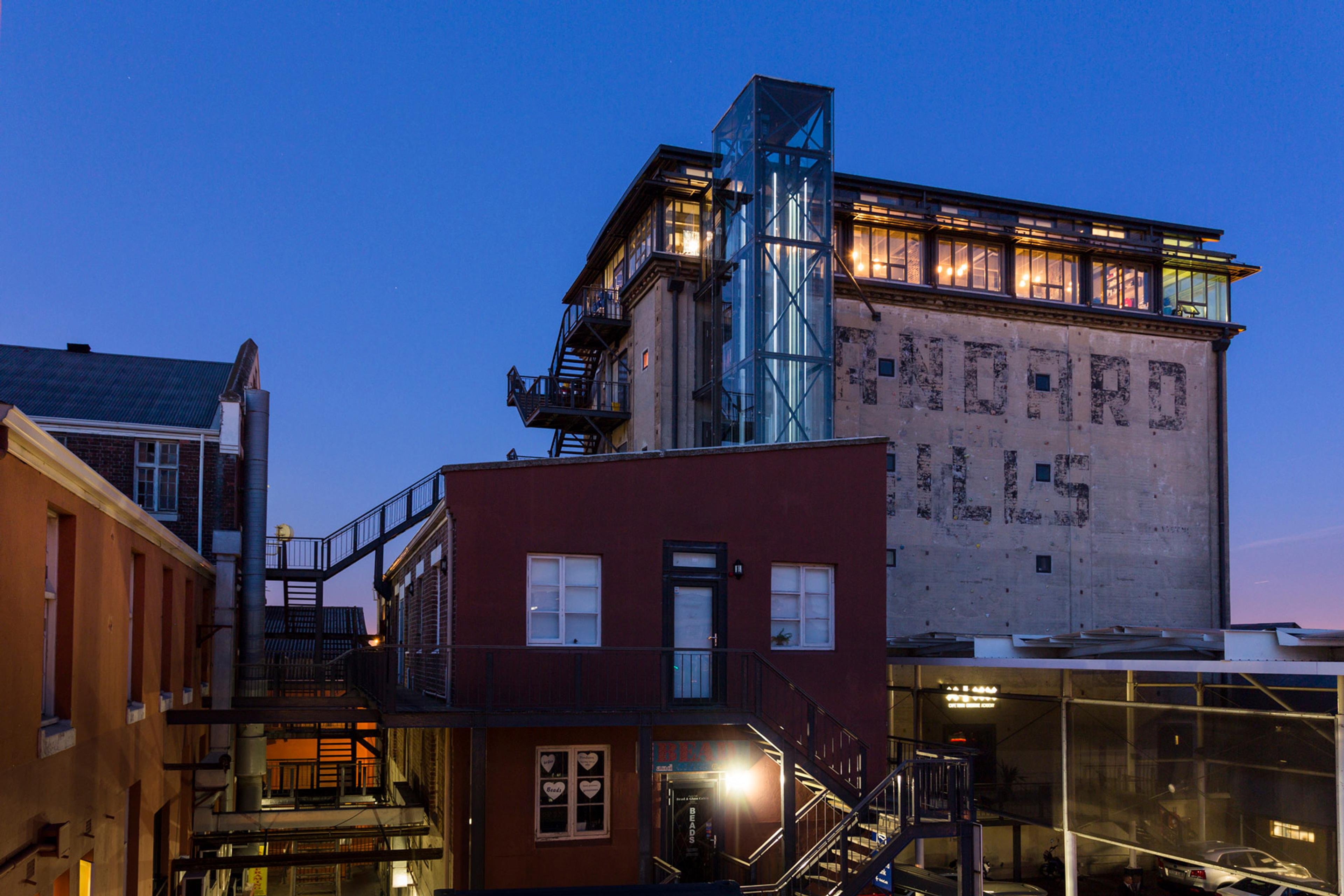 The old mill silo at dusk, its glazed top floor lit, above the brick buildings of the Old Biscuit Mill