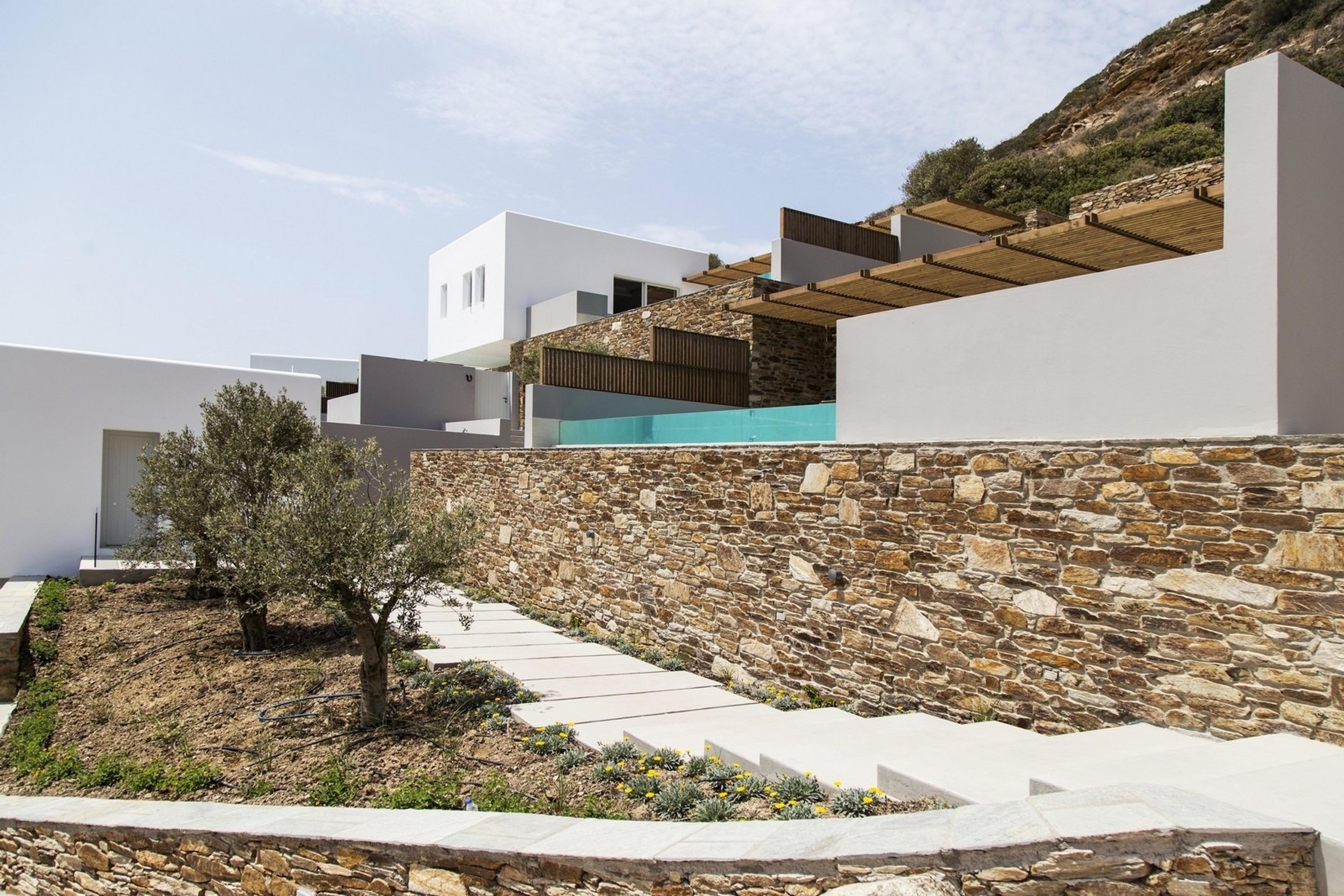 A stone retaining wall climbs a hillside, with steps and olive trees below and white building blocks with timber pergolas above.