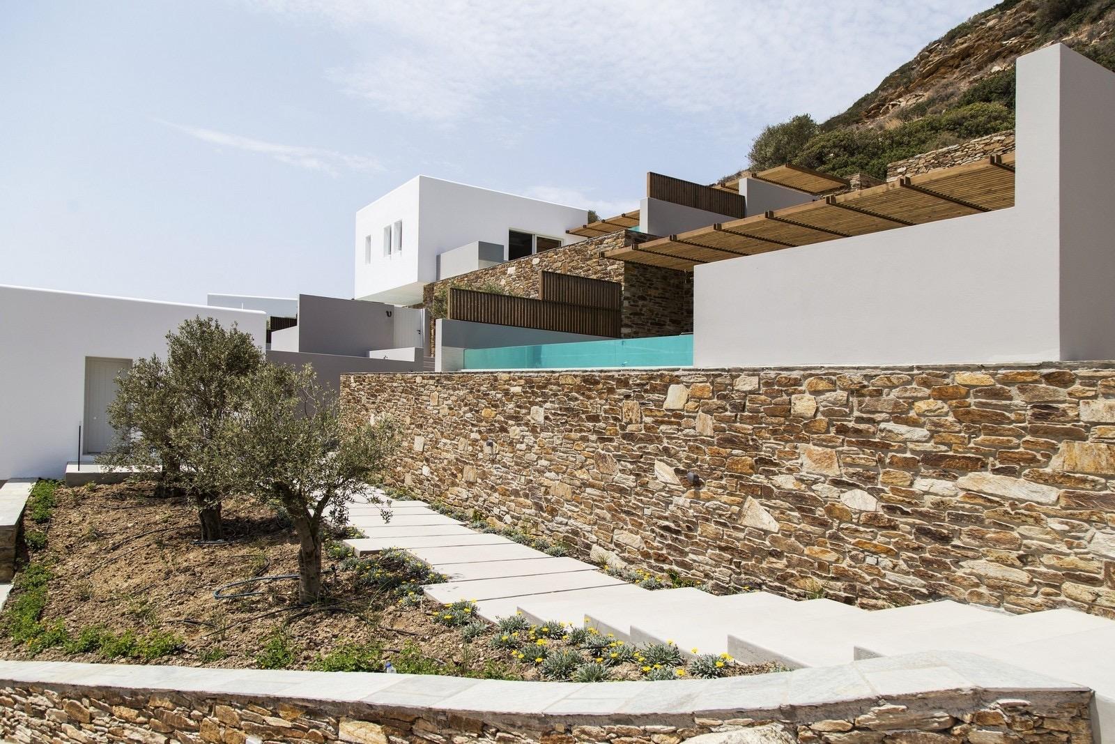A stone retaining wall climbs a hillside, with steps and olive trees below and white building blocks with timber pergolas above.