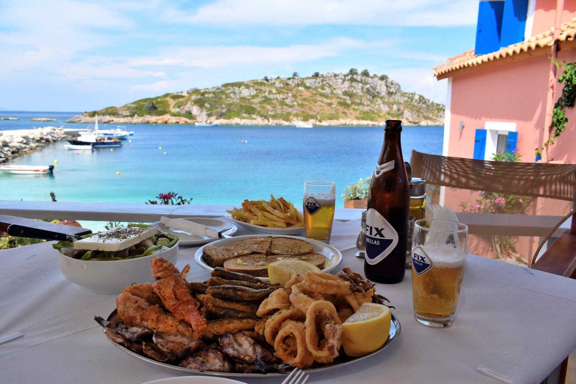 A table of fried fish, calamari, Greek salad, chips and beer on a terrace above the harbour at Agios Nikolaos, with boats moored and the islet beyond