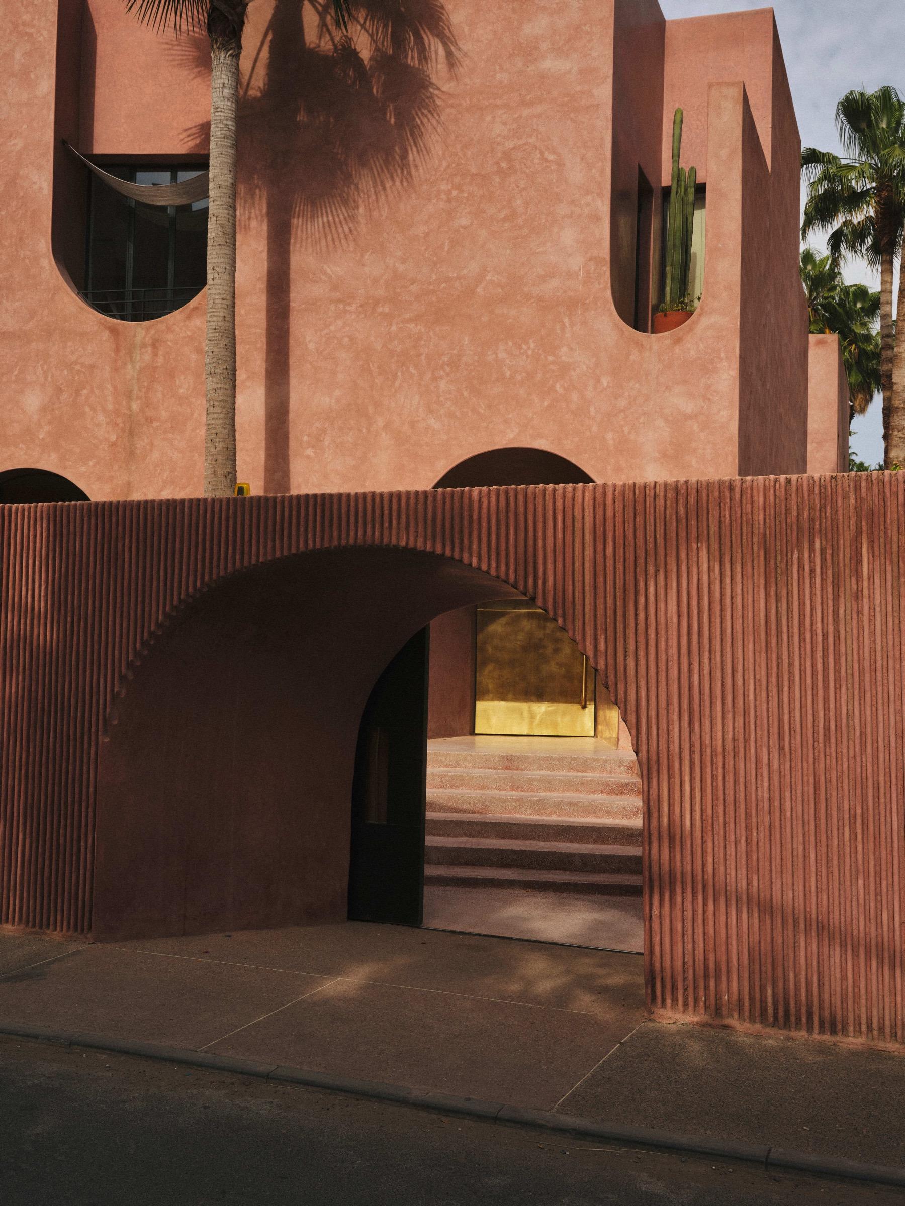 a pink stuccoed building facade with an arched entrance passage leading to a lit golden door and steps, palm trees behind
