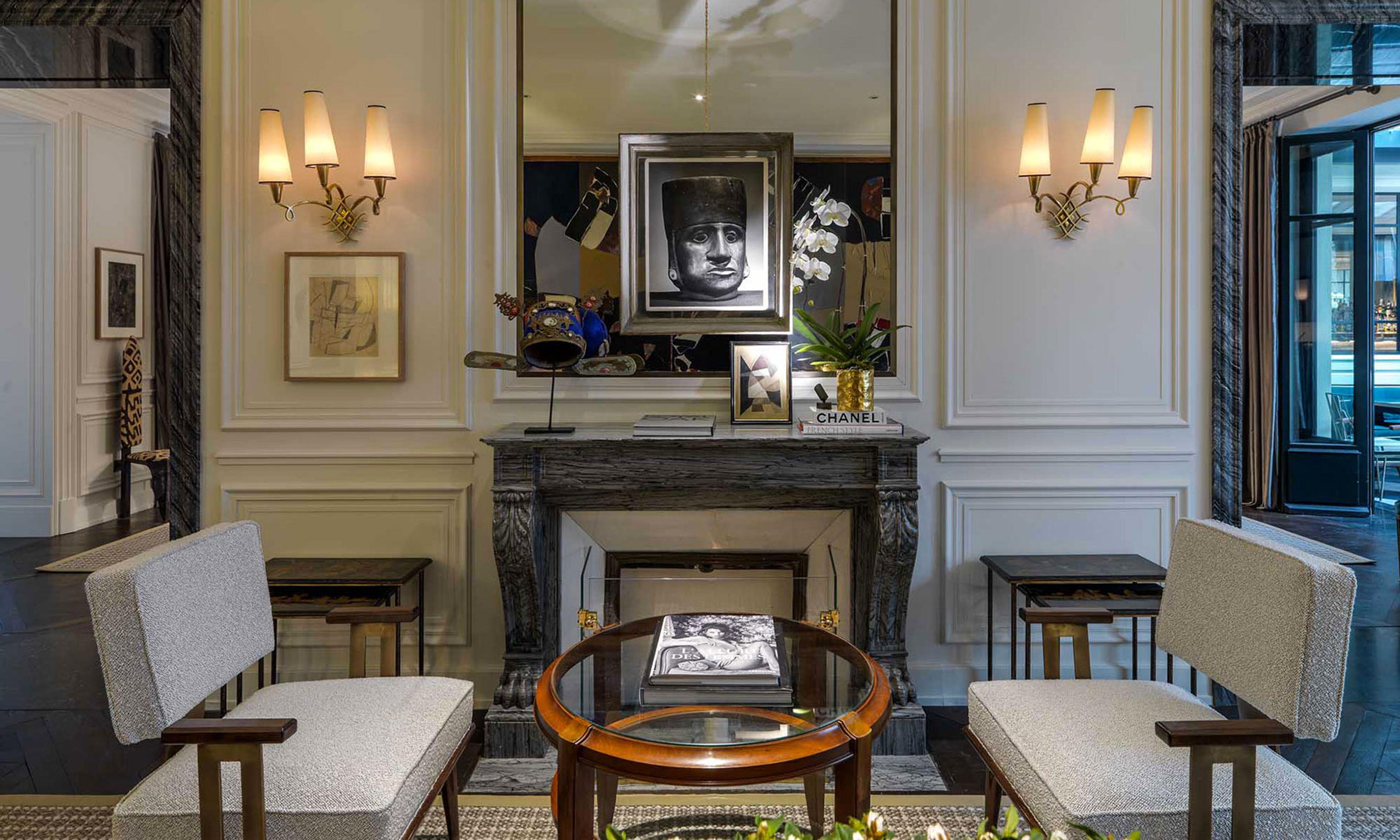 A sitting room with twin wall sconces either side of a mirror, an orchid and framed artwork on a marble mantelpiece, and two upholstered armchairs around a low table.