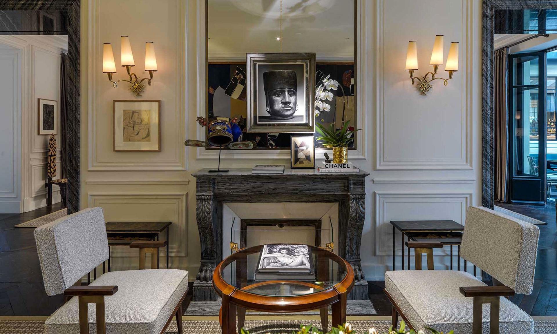 A sitting room with twin wall sconces either side of a mirror, an orchid and framed artwork on a marble mantelpiece, and two upholstered armchairs around a low table.