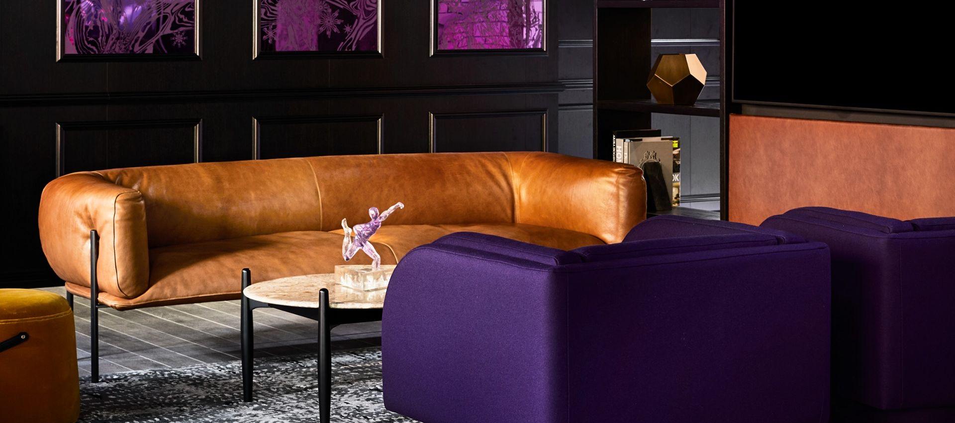 a lounge with a curved tan leather sofa, a deep purple armchair, and framed artwork on a dark panelled wall