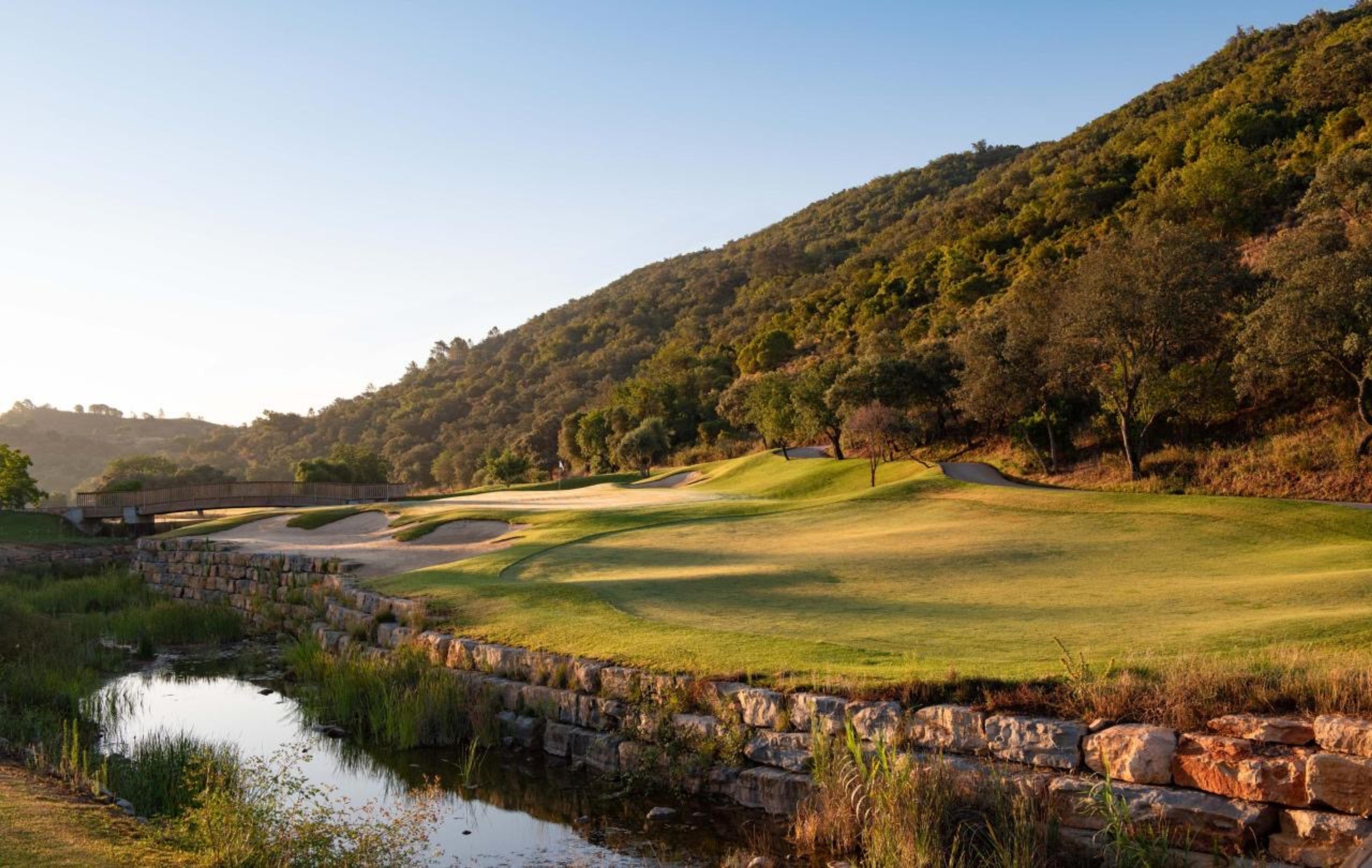 A golf course green beside a stream and a stone retaining wall, with a wooden footbridge and wooded hills behind.