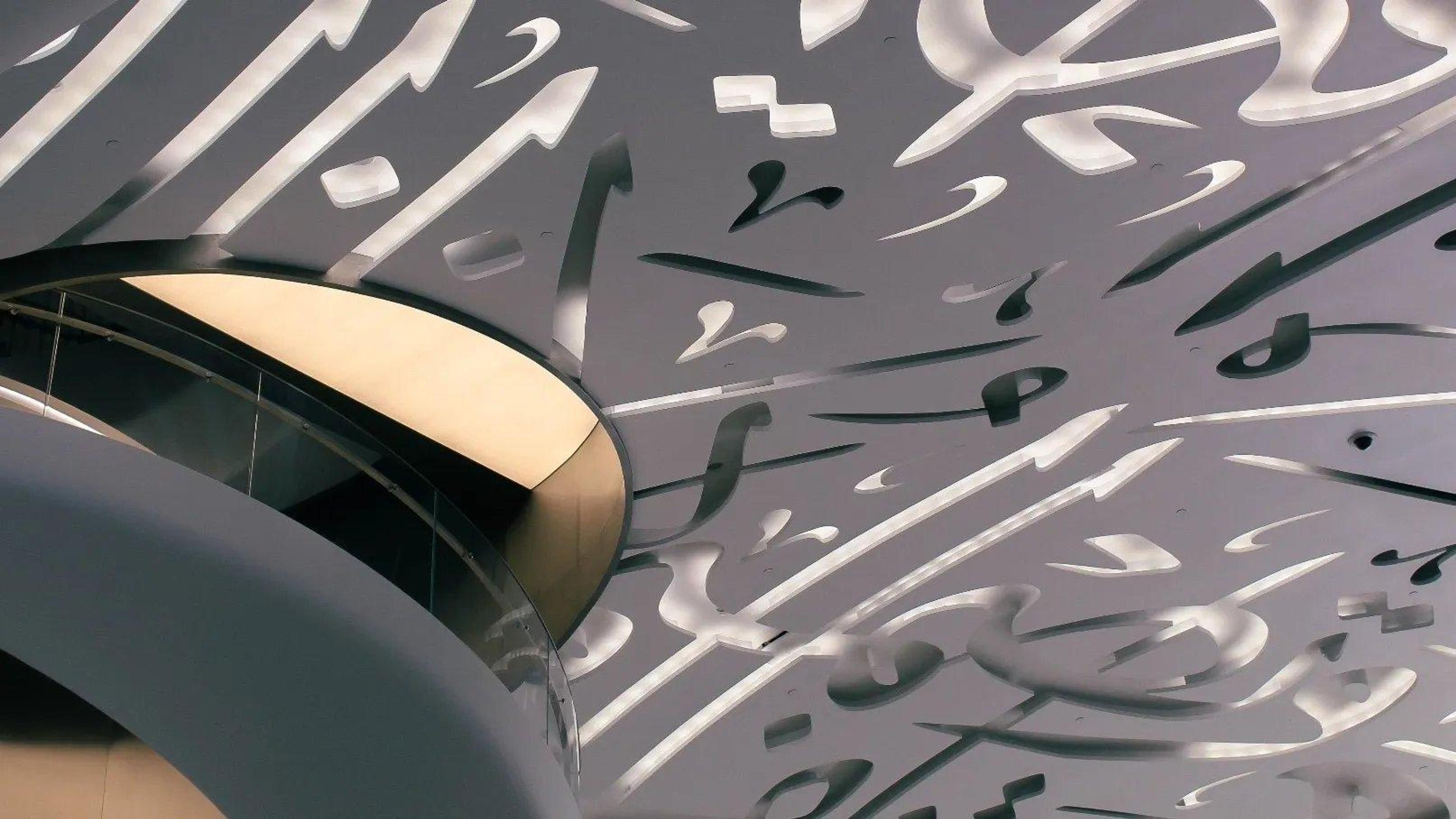 A close view of a ceiling and balcony covered in cut-metal Arabic calligraphy, with a curved mezzanine edge lit from below.