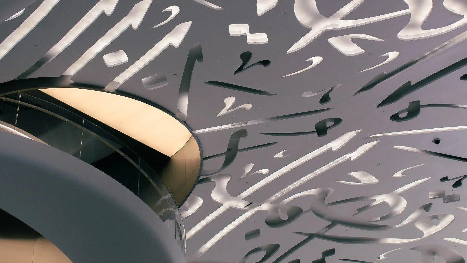 A close view of a ceiling and balcony covered in cut-metal Arabic calligraphy, with a curved mezzanine edge lit from below.