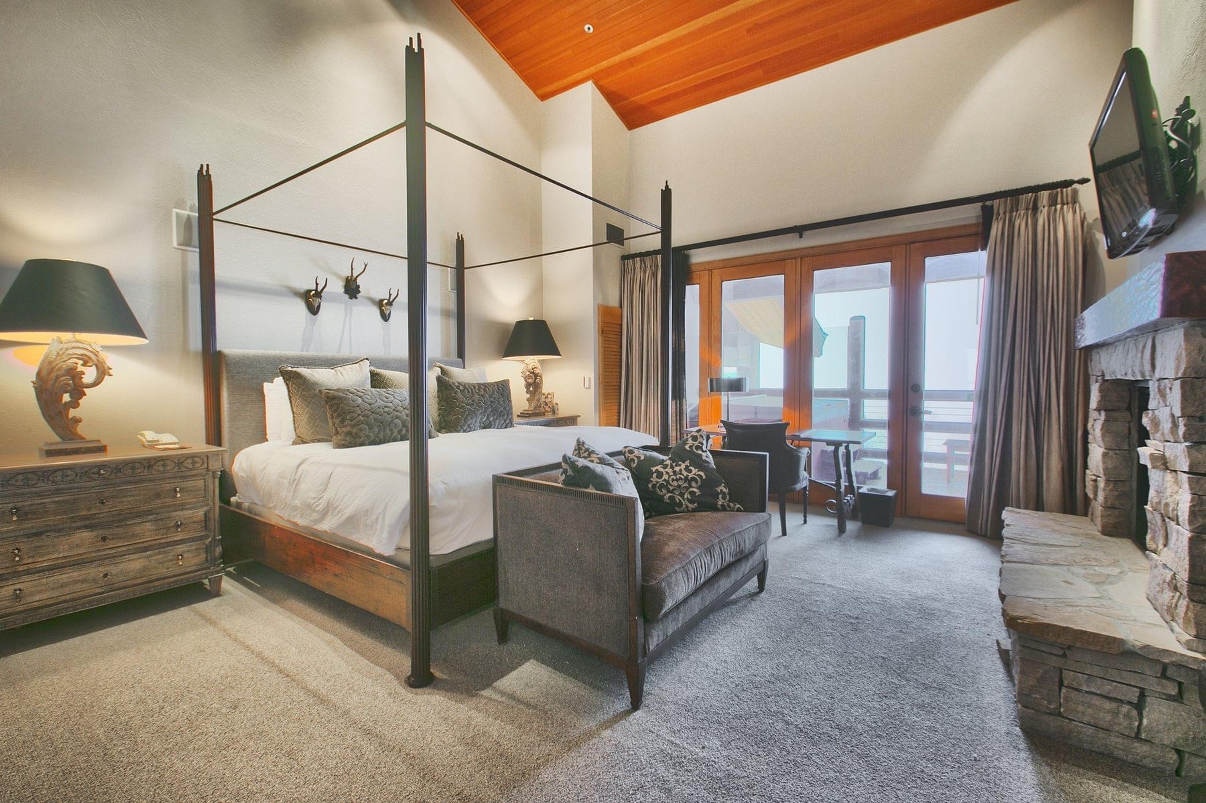 a bedroom with a dark four poster canopy bed, mounted antlers above the headboard, a stone fireplace, and glass doors onto a balcony