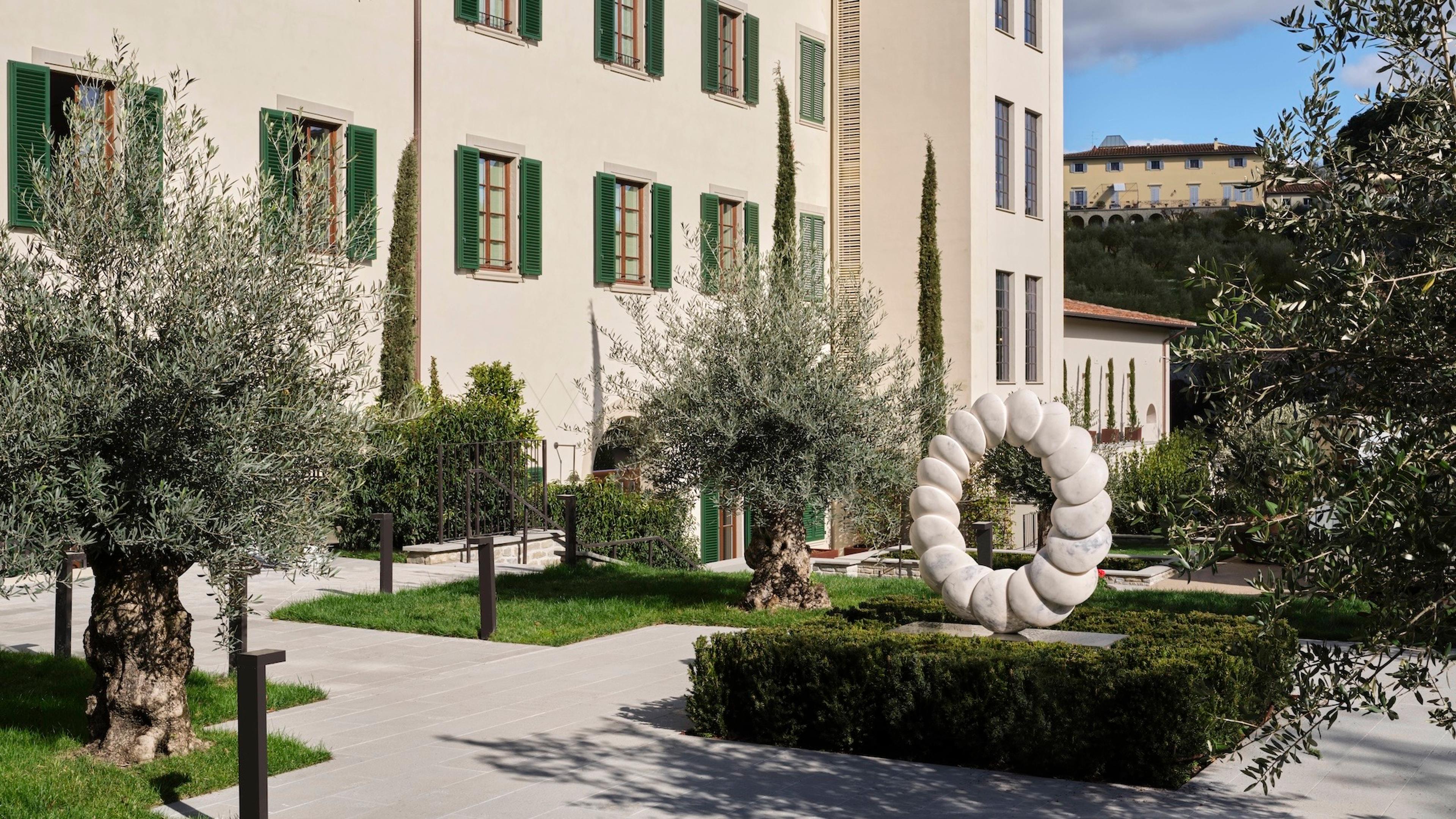 a courtyard of mature olive trees and a coiled white marble sculpture in front of a cream building with green shutters