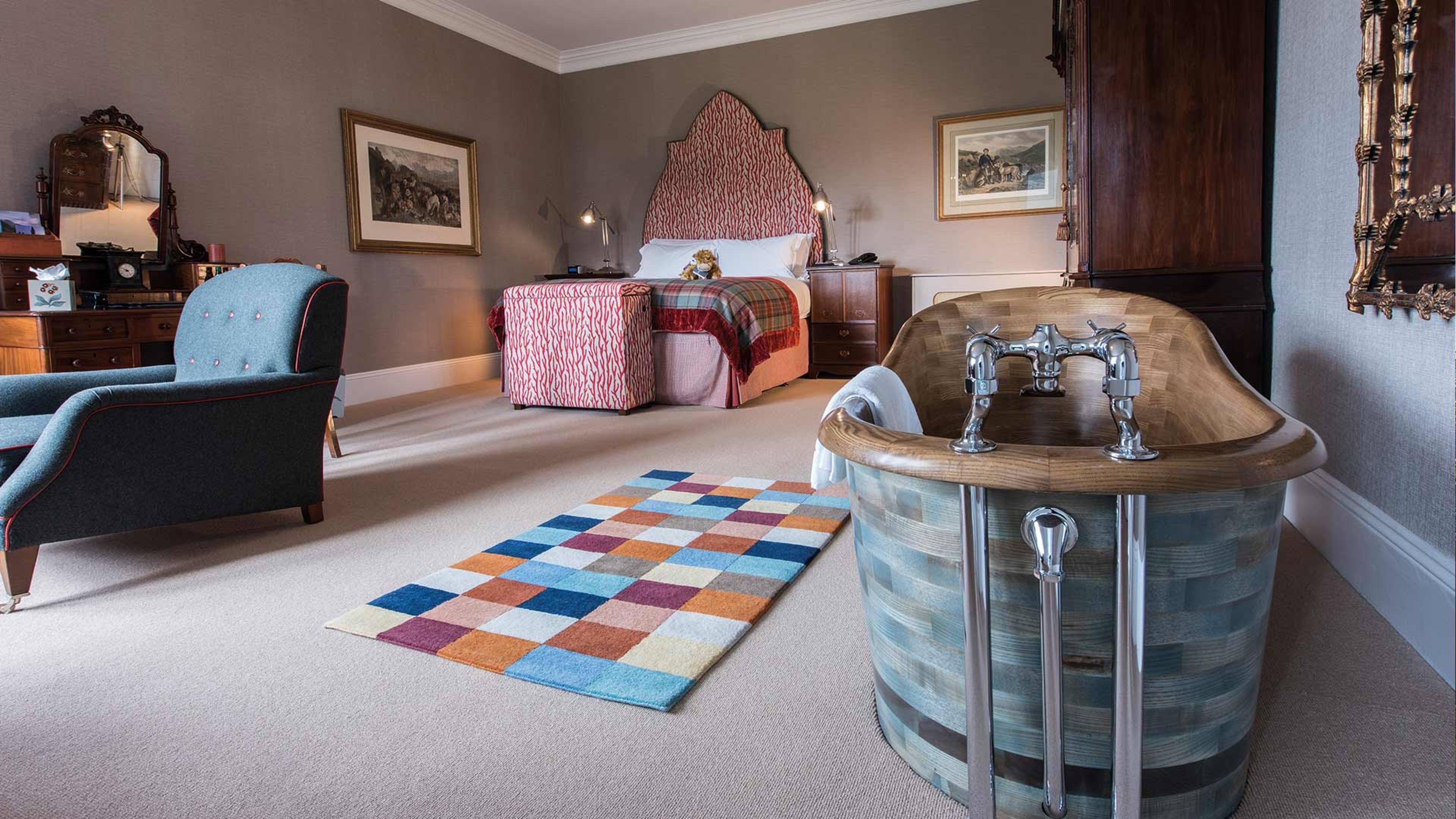 A hotel room with a free standing wood and copper slatted bathtub in the foreground, a patterned headboard and a blue armchair beyond, on a patchwork coloured rug.