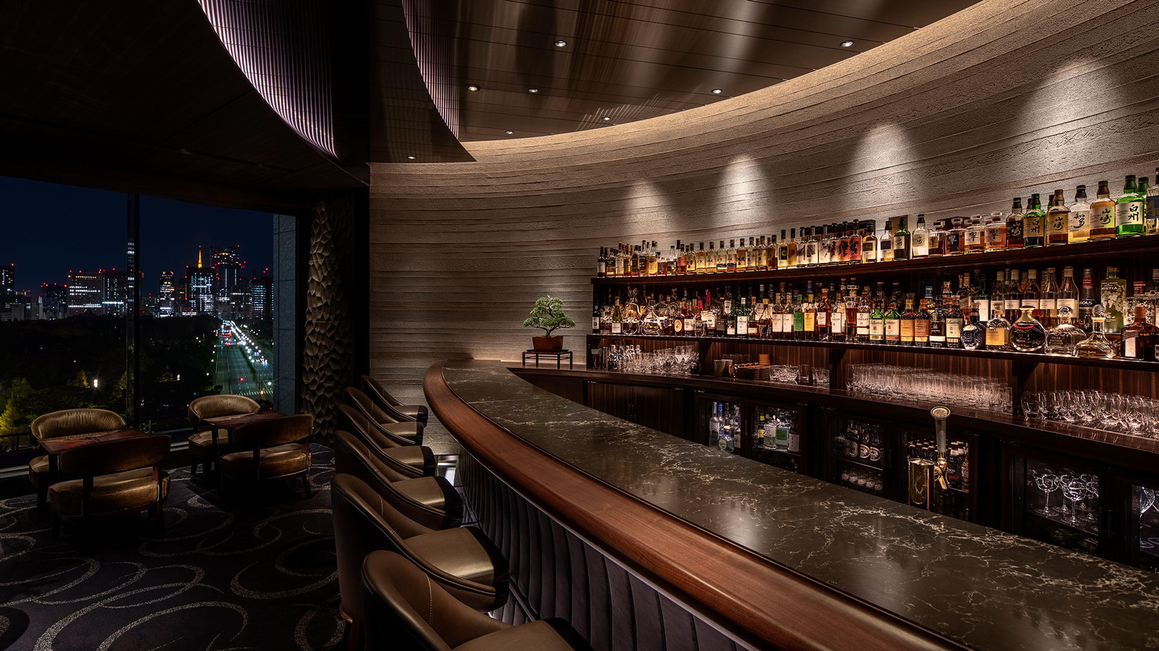 A curved hotel bar at night, shelves of spirits lit along the wall, leather stools at the counter and the city skyline through the window.